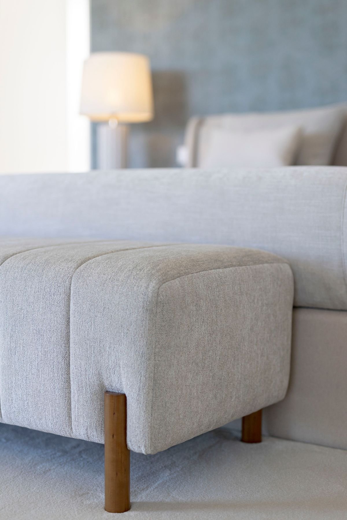 Detalhe aproximado dos pés em madeira e do estofo acolchoado da banqueta disposta junto à cama.