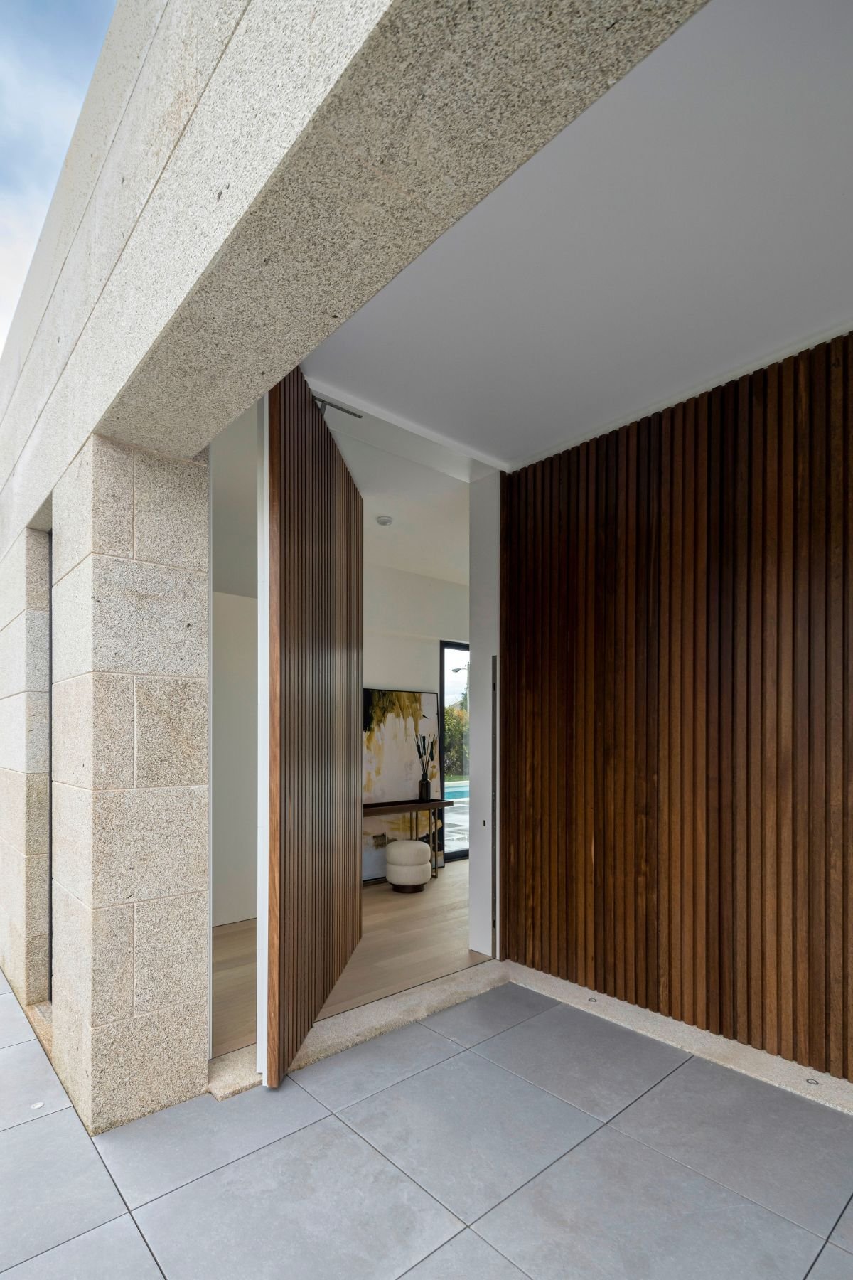 Porta pivotante ripada e fachada em pedra com vista para hall de entrada pequeno e consola entrada, refletindo a harmonia de interior design à entrada.