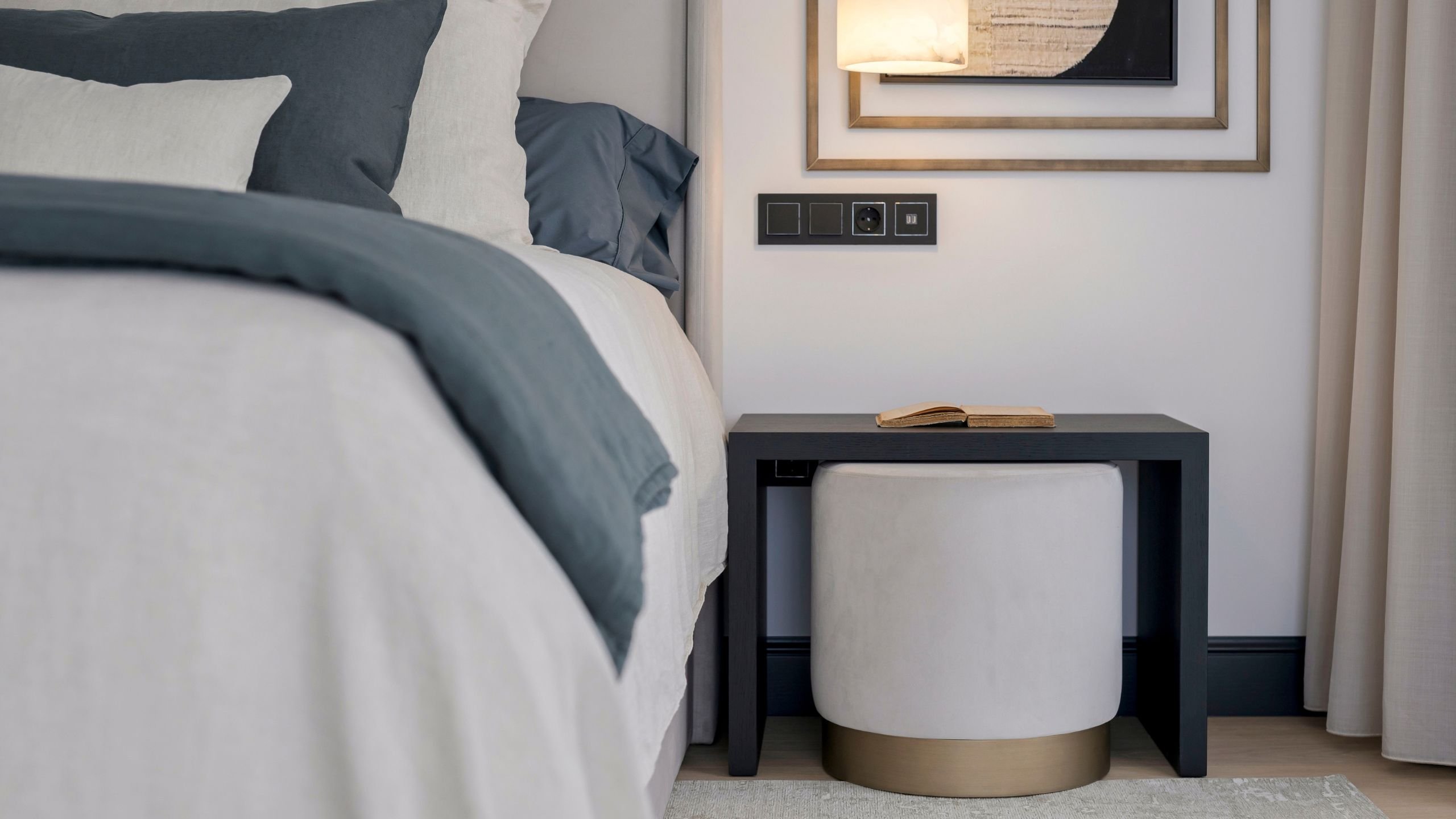Lateral de cama de casal com mesa de cabeceira preta e puff cilíndrico encaixado na decoração de quarto.