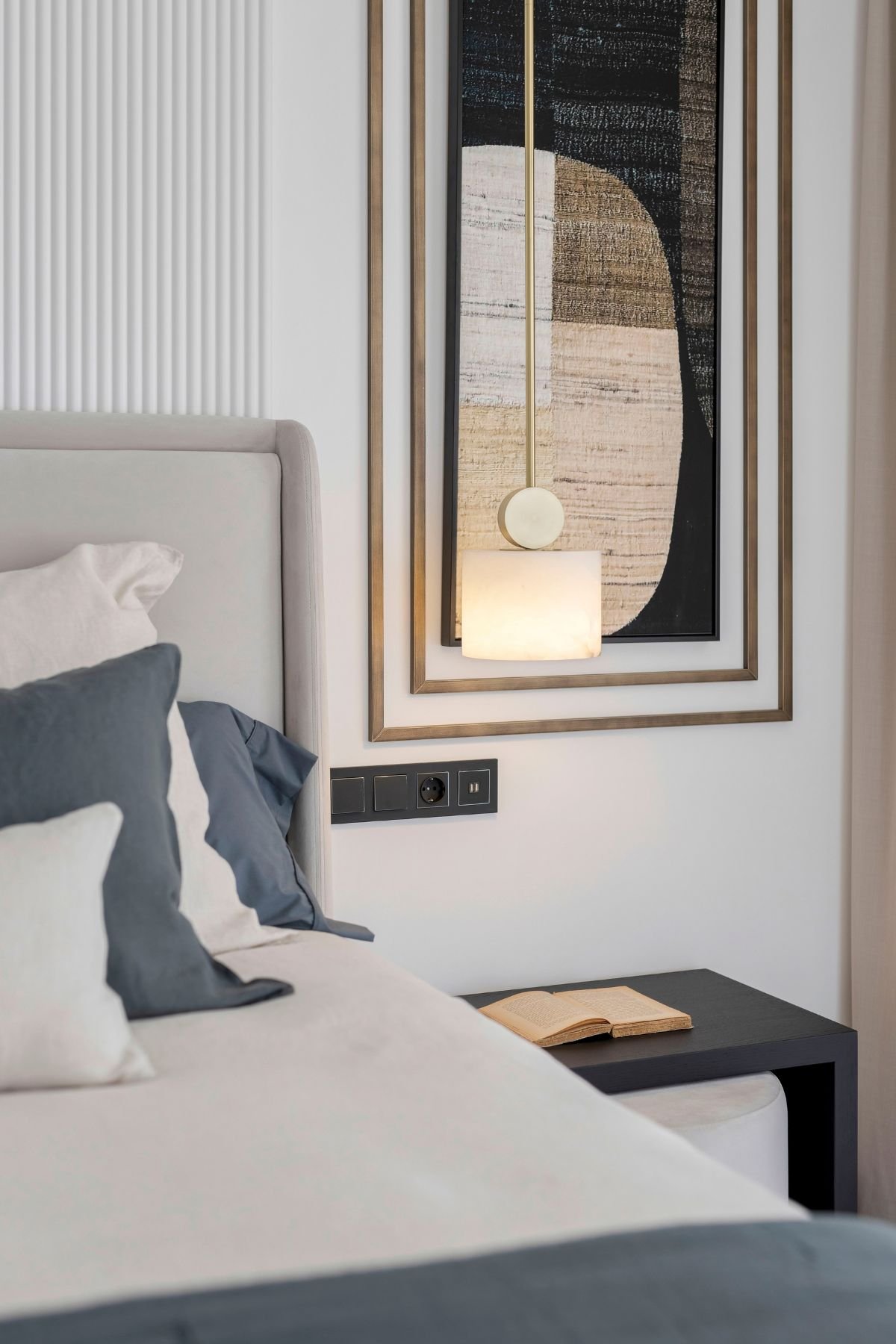 Cama de casal estofada ao lado de mesa de cabeceira preta e candeeiro suspenso em elegante decoração de quarto.