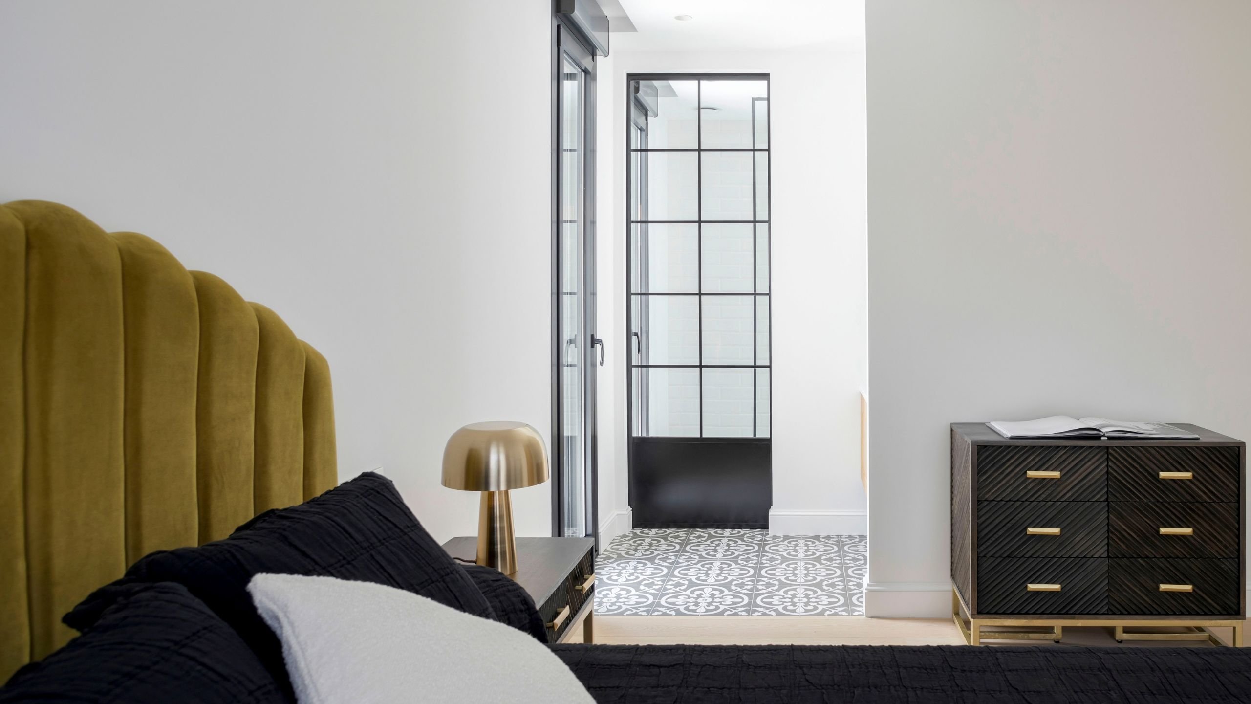 Vista de quarto com cama de casal de cabeceira mostarda e passagem para a suite, refletindo um requintado design de interiores.