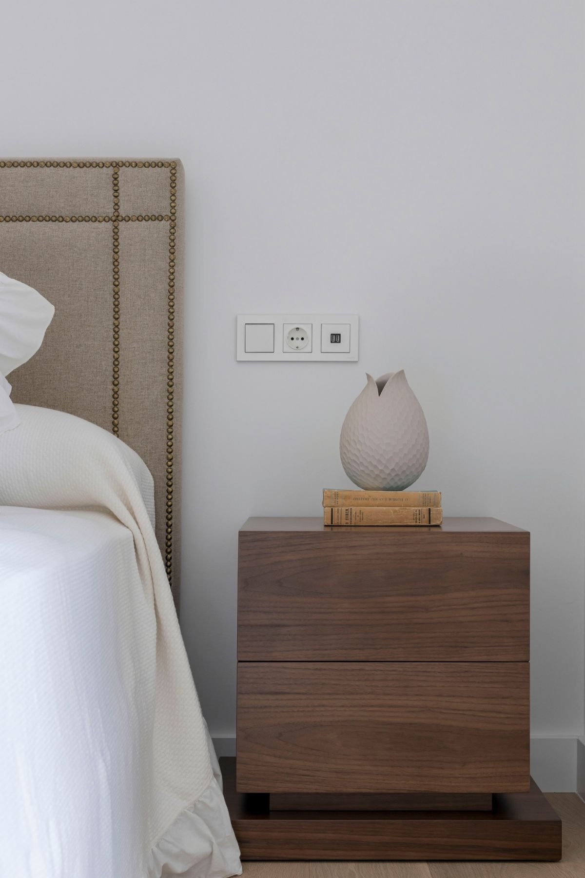 Mesa de cabeceira em madeira com jarra decorativa e livros junto à cama em quarto de hóspedes, destacando o requinte do design de interiores na decoração de quarto.