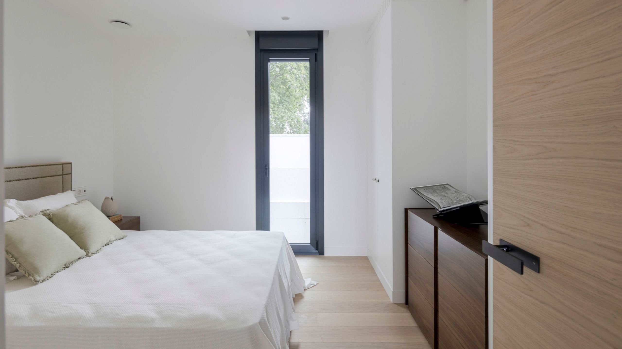Cama de casal com roupa de cama branca e cómoda em madeira num quarto de hóspedes, evidenciando o design de interiores e a decoração de quarto elegante.