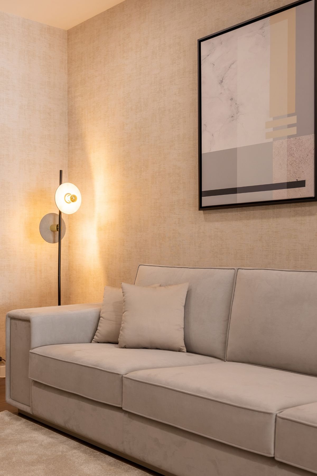 Pormenor do confortável sofá bege sob a iluminação acolhedora do candeeiro de pé e quadros decorativos.
