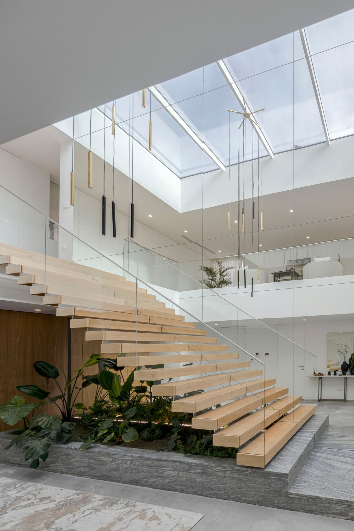 Vista ampla da arquitetura interior com escada em consola de madeira, claraboia no teto, lustre moderno suspenso e jardim interior verdejante.
