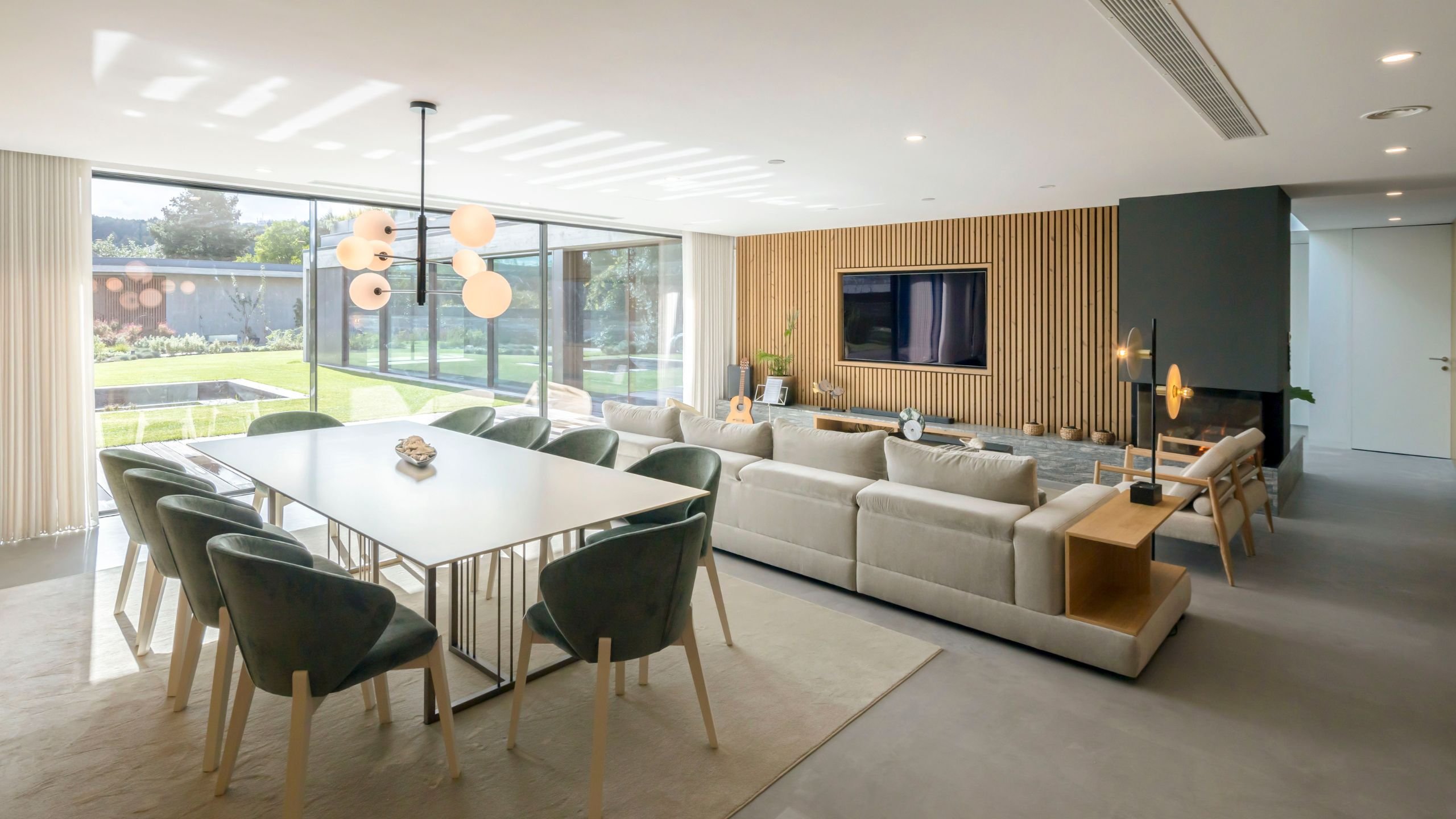 Sala de estar e jantar em open space com sofá claro, mesa de jantar para oito pessoas, cadeiras em tecido verde e painel de ripas de madeira com televisão.