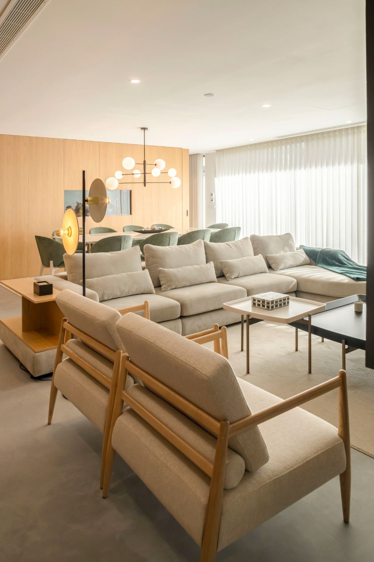 Perspetiva da sala de estar com sofá modular beige, cadeirões em madeira clara, mesas de centro e candeeiro de pé contemporâneo junto à área de refeições.
