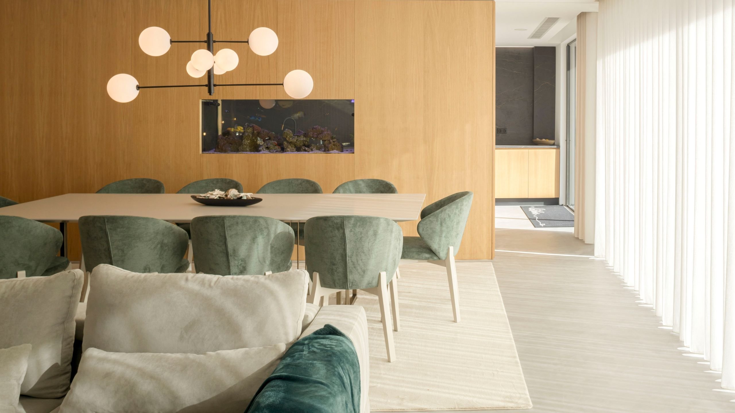 Zona de jantar com mesa para oito pessoas, cadeiras verdes e aquário encastrado em painel de madeira natural sob iluminação de candeeiro moderno.