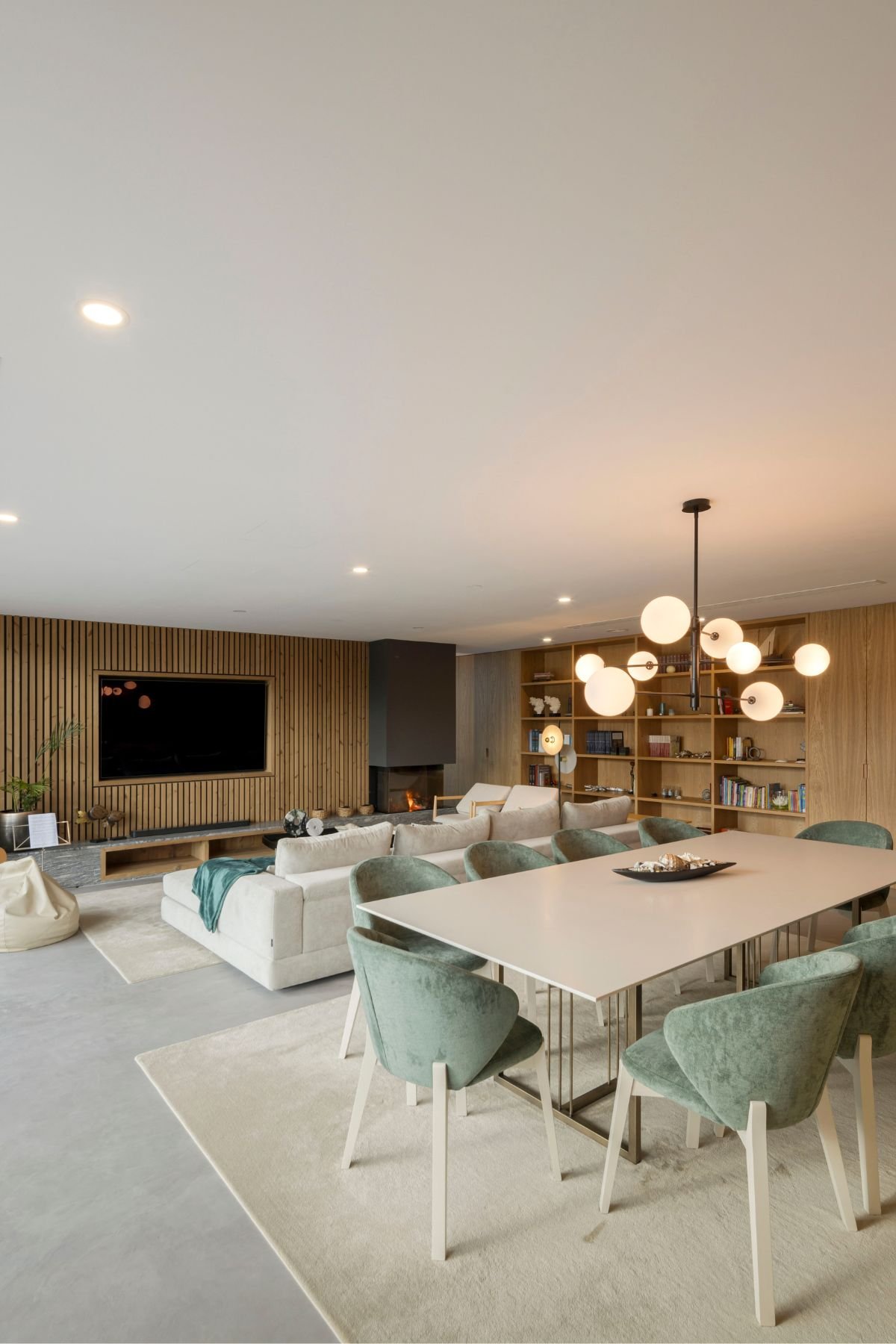 Vista geral do espaço open space integrando sala de jantar com mesa e cadeiras verdes, e sala de estar com sofá e painel de TV em ripas de madeira.