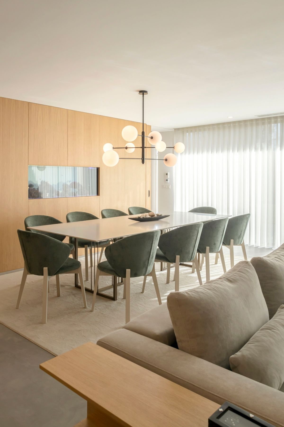 Mesa de jantar com tampo claro sobre tapete neutro, rodeada por cadeiras estofadas em verde, junto a cortinas brancas translúcidas.