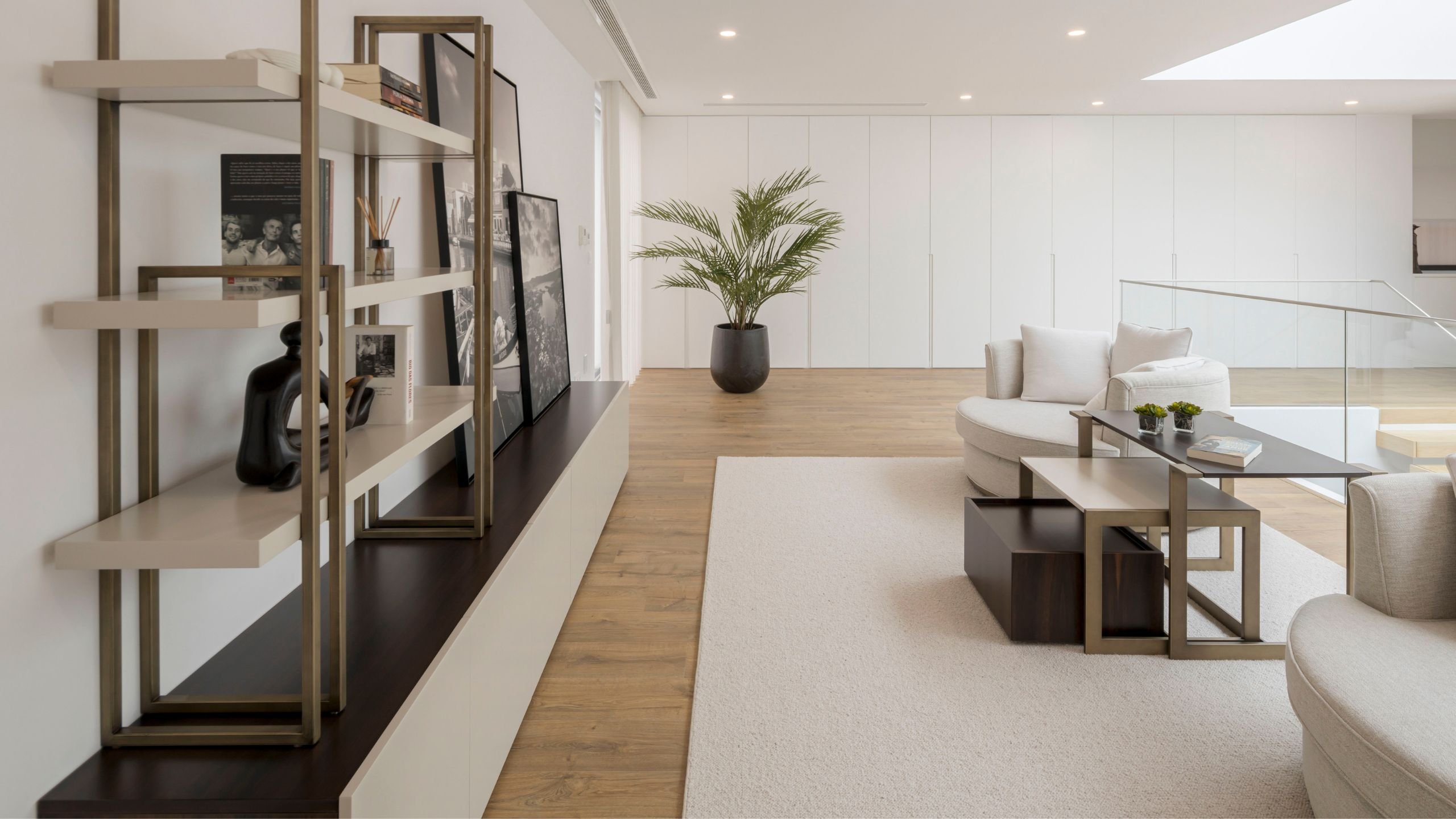 Vista ampla da área de descanso com estante de livros, móvel baixo com quadros decorativos, tapete claro e cadeirões dispostos em torno da mesa de apoio.