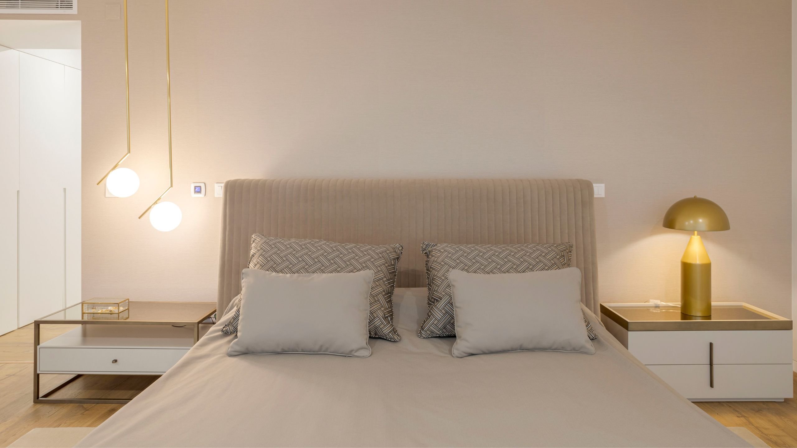 Cama com cabeceira estofada em tom bege canelado, ladeada pelas mesas de cabeceira com iluminação suspensa moderna em metal dourado e candeeiro de mesa dourado.