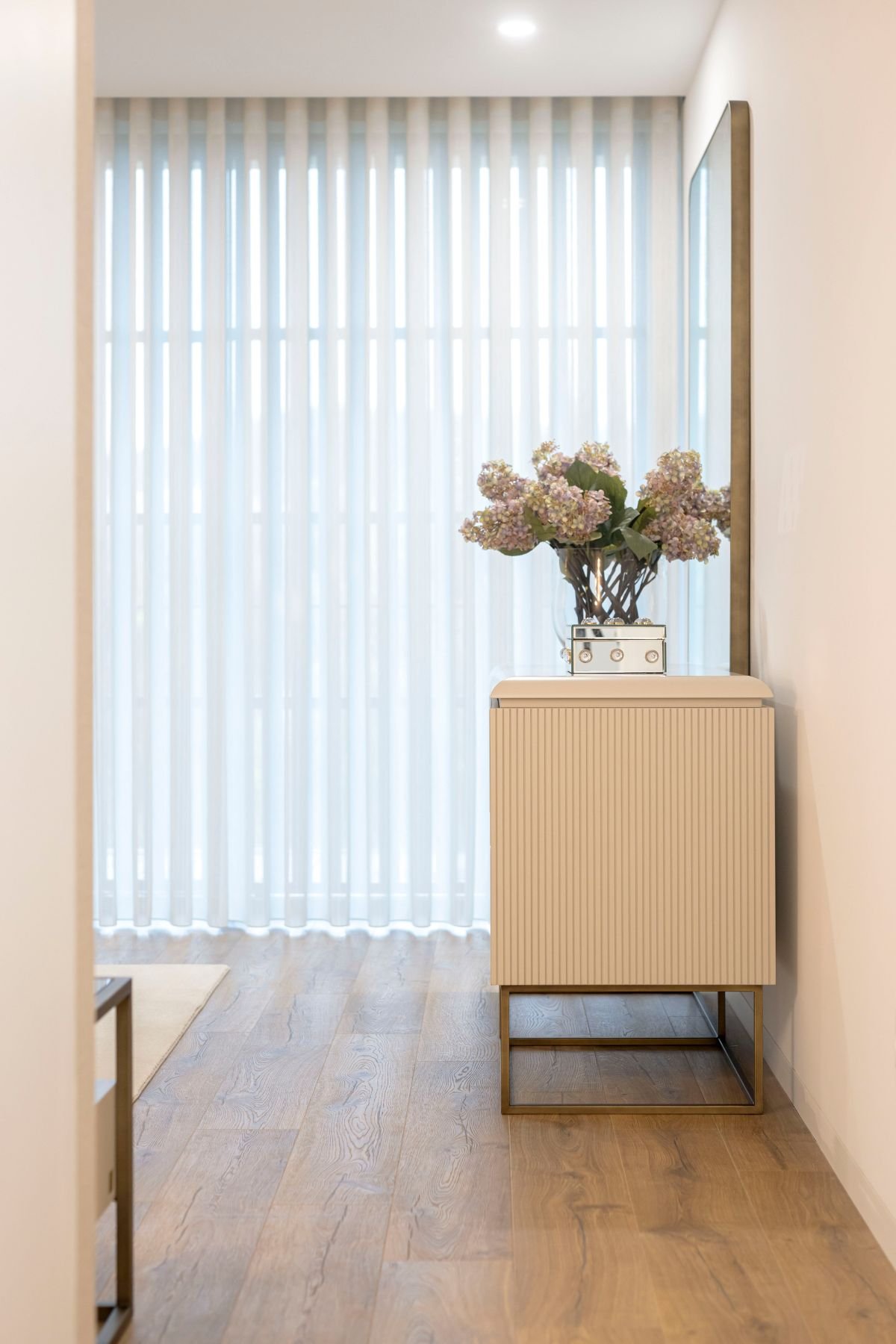 Pormenor lateral da cómoda com acabamento canelado e pés dourados, decorada com vaso de hortênsias e posicionada junto a cortinas brancas translúcidas.