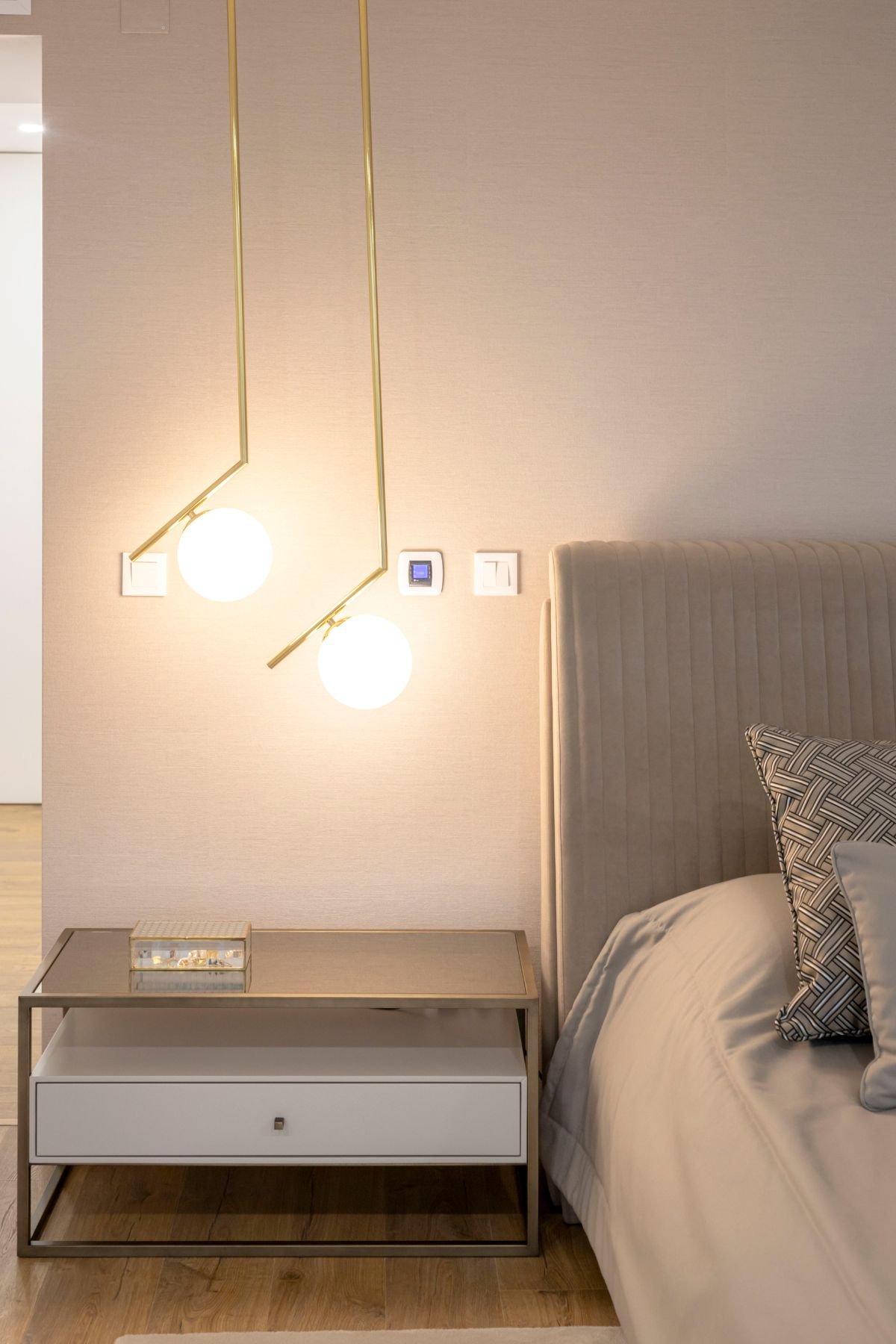 Detalhe da mesa de cabeceira com gaveta branca e estrutura metálica, sob a luz quente de dois candeeiros suspensos de vidro esférico e haste dourada.