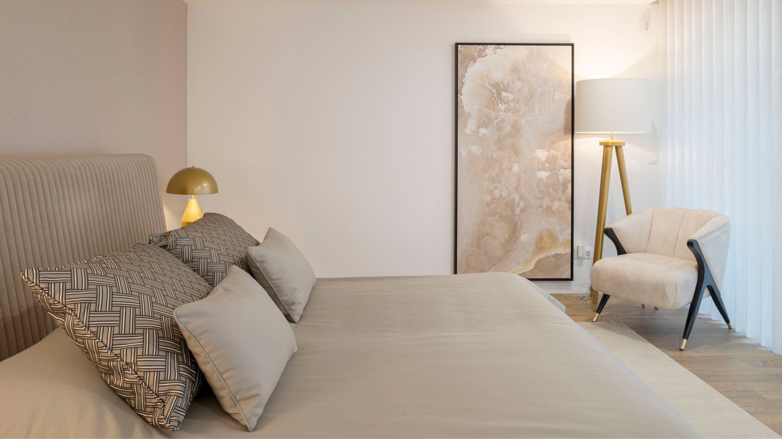 Perspetiva do quarto destacando a cama em tons neutros, cadeirão de leitura ao fundo com pés pretos e dourados, candeeiro de pé alto em tripé e painel decorativo na parede.