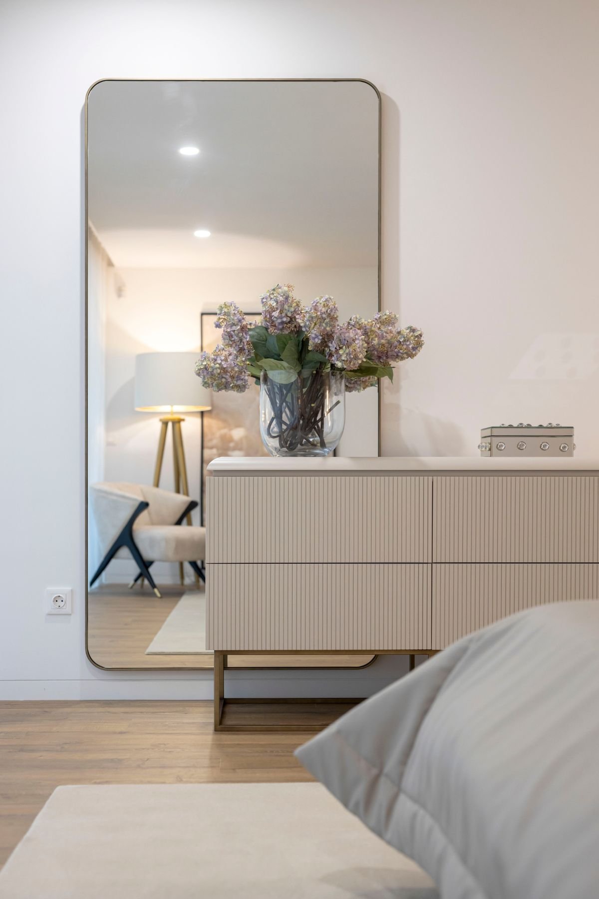 Cómoda canelada em tom neutro com vaso de flores sobre o tampo, ladeada pelo espelho de corpo inteiro de cantos arredondados que reflete a zona de leitura do quarto.