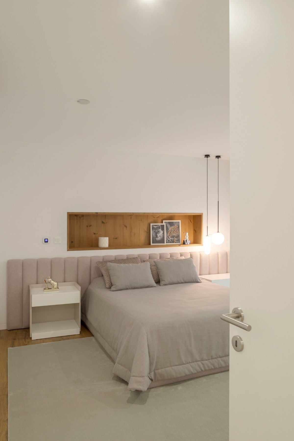 Perspetiva ampla do quarto com a camade cabeceira estofada rosa-pálido, nicho em madeira com fotos, iluminação suspensa moderna e mesa de cabeceira branca.