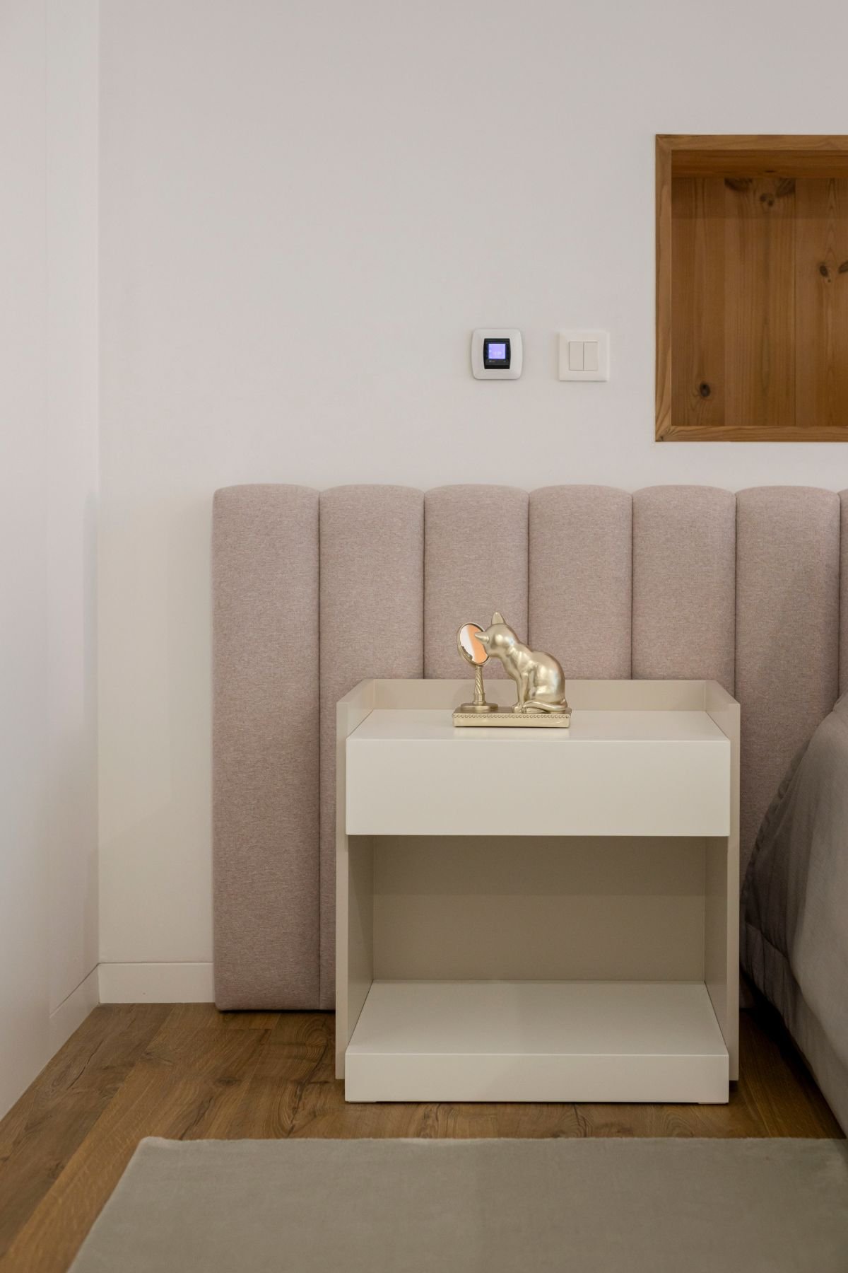 Pormenor da mesa de cabeceira branca acompanhada por uma figura decorativa dourada em forma de gato junto à cabeceira estofada de módulos verticais.