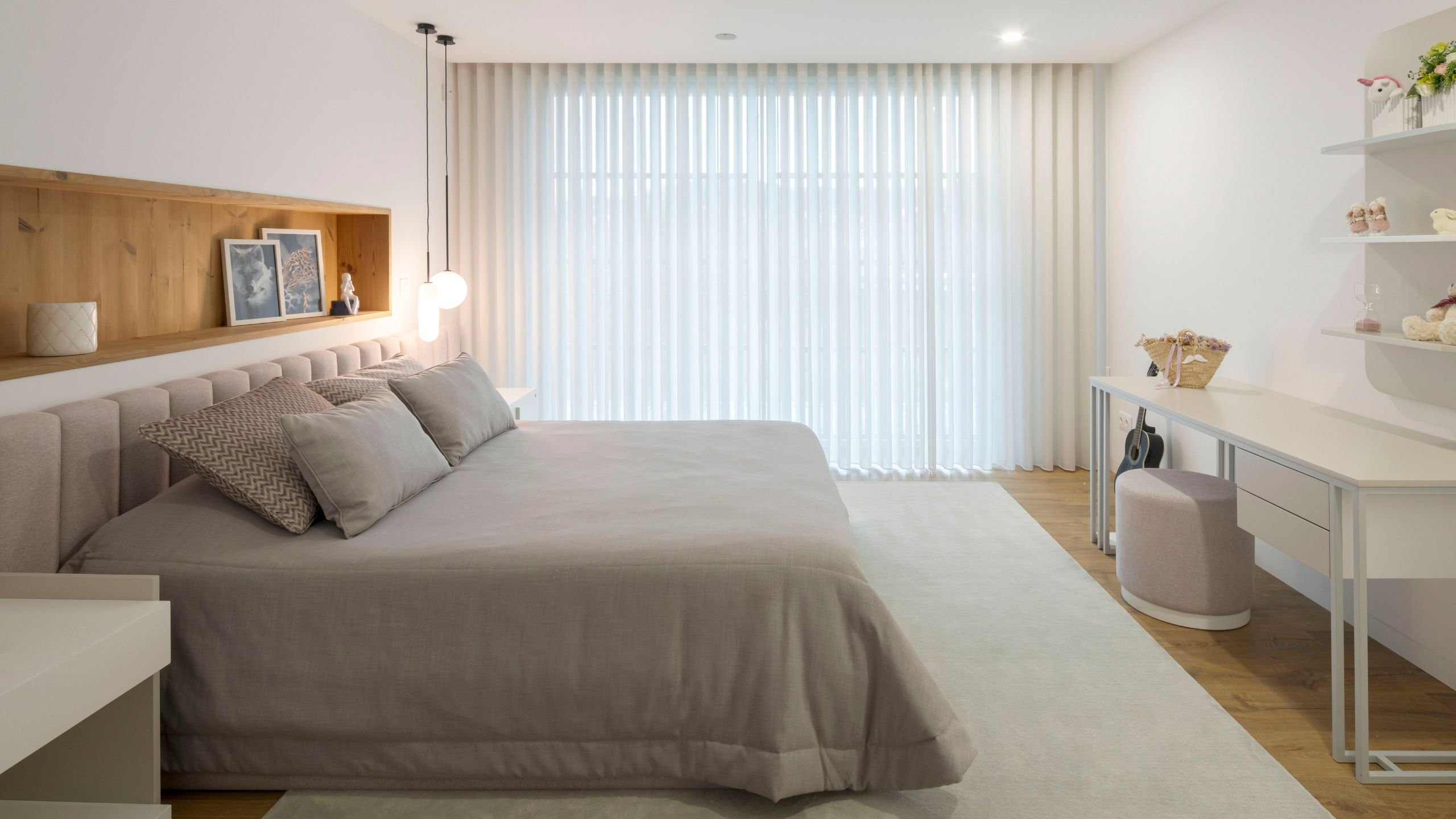 Vista geral da suite destacando a cama em tons neutros, grandes cortinados brancos, área de estudo com secretária, pufe estofado e prateleiras decorativas.