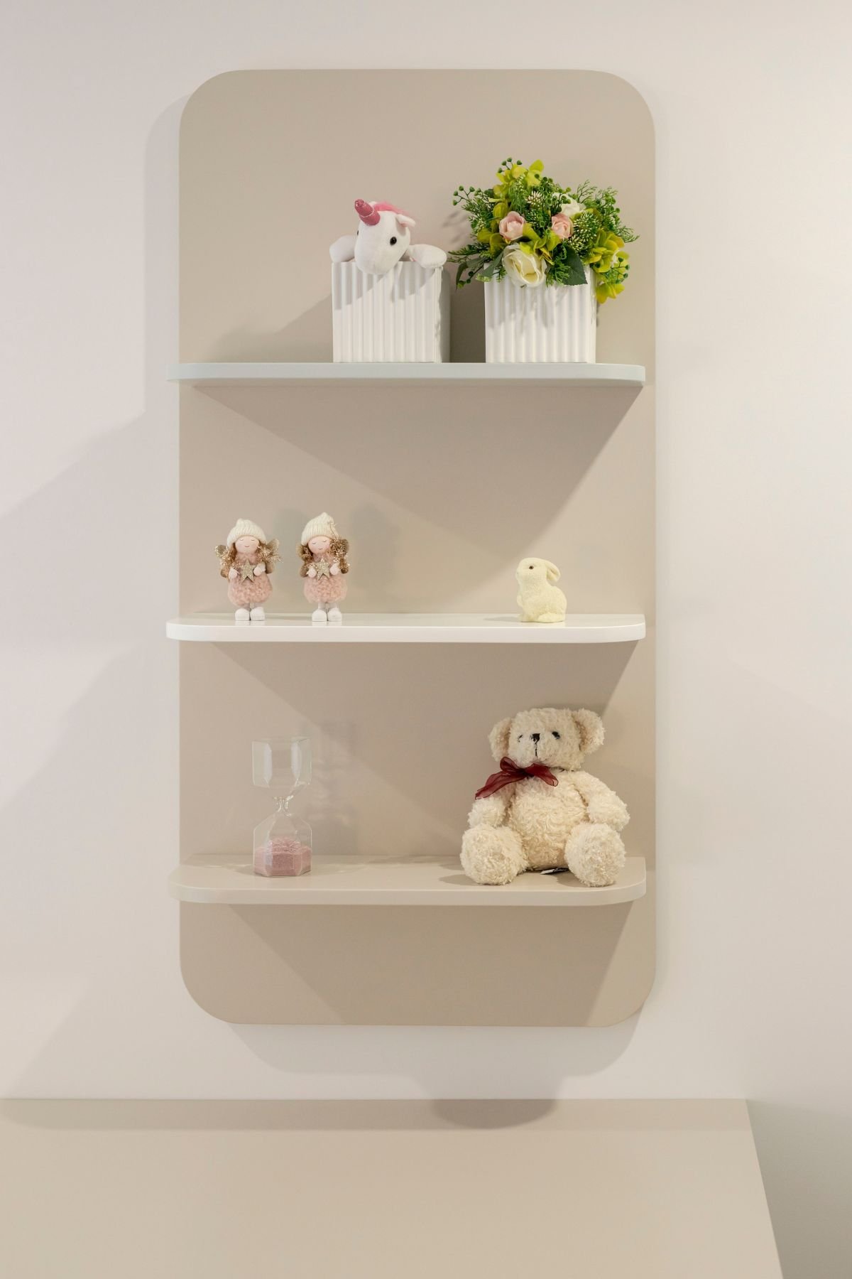 Painel de parede bege com prateleiras arredondadas decoradas com peluches, arranjos florais e pequenos objetos ornamentais.