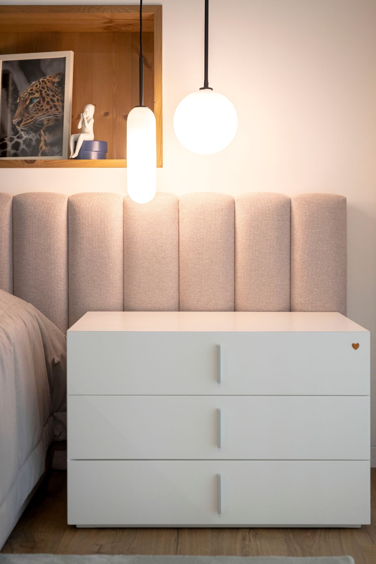 Pormenor da mesinha de cabeceira branco de três gavetas ao lado da cama, sob a luz difusa de dois candeeiros suspensos.