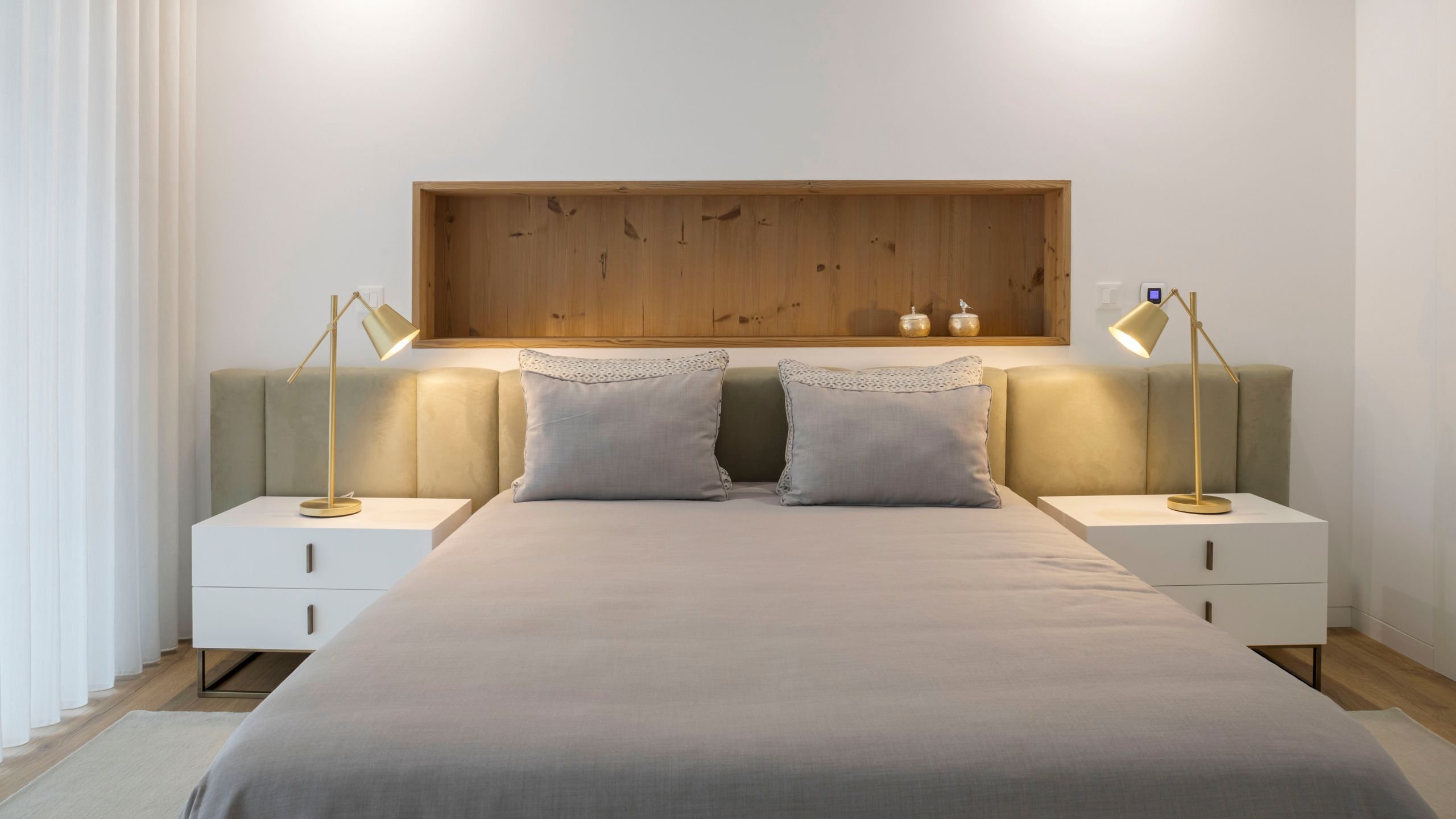 Perspetiva frontal do quarto de hóspedes com cama de cabeceira estofada em verde-oliva, mesas de cabeceira brancas, candeeiros de mesa articulados dourados e nicho em madeira na parede.