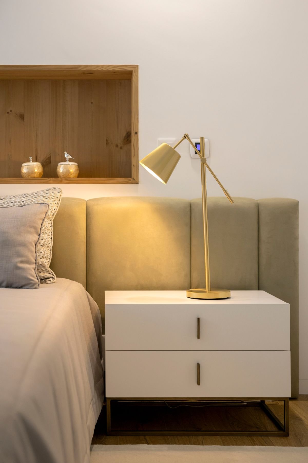Pormenor da mesa de cabeceira branca com gavetas e base metálica escura, iluminada por candeeiro de mesa articulado dourado junto à cabeceira em tom verde.