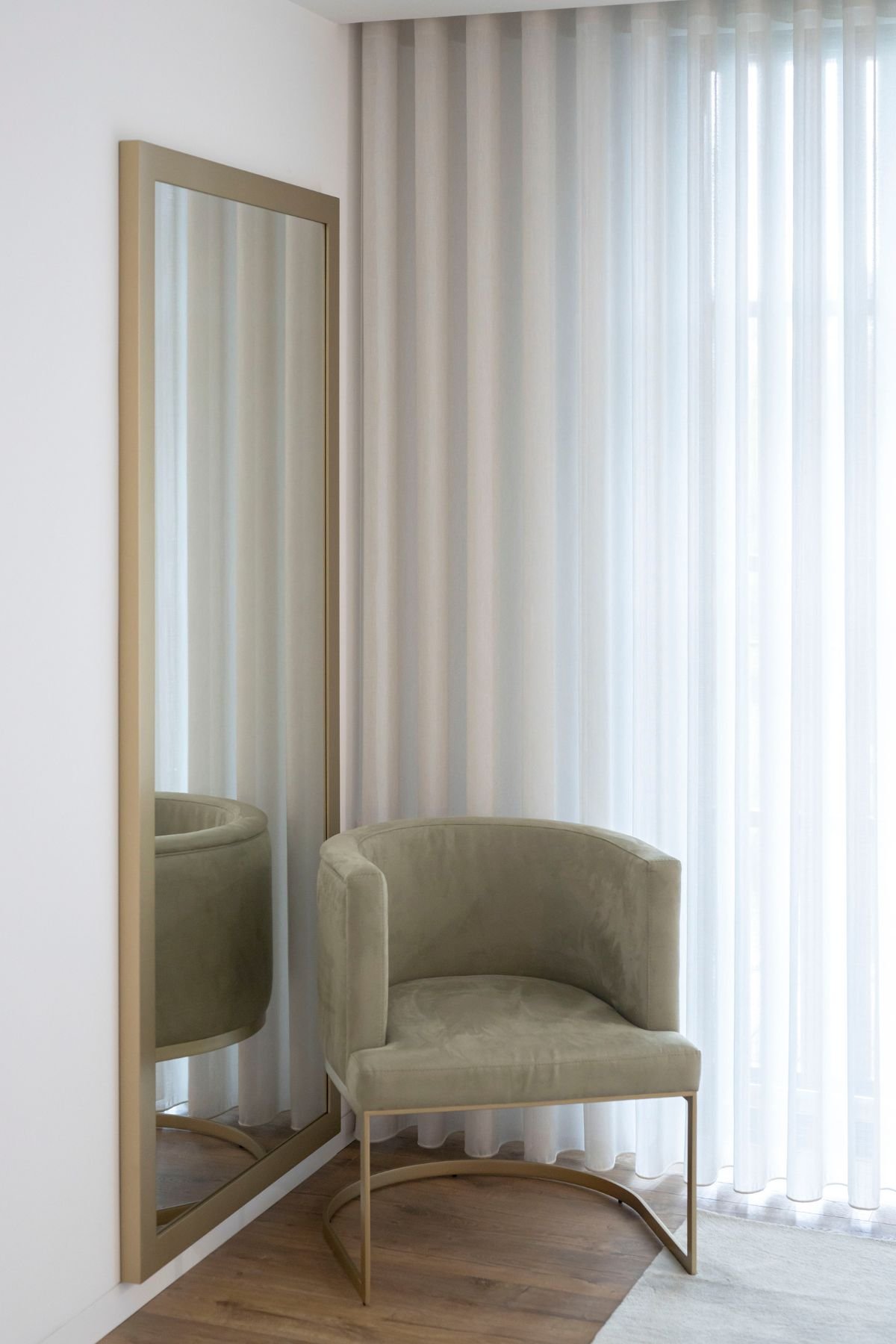 Cadeirão de apoio estofado em veludo verde com estrutura metálica dourada, posicionado junto a espelho retangular de corpo inteiro e cortinas brancas.