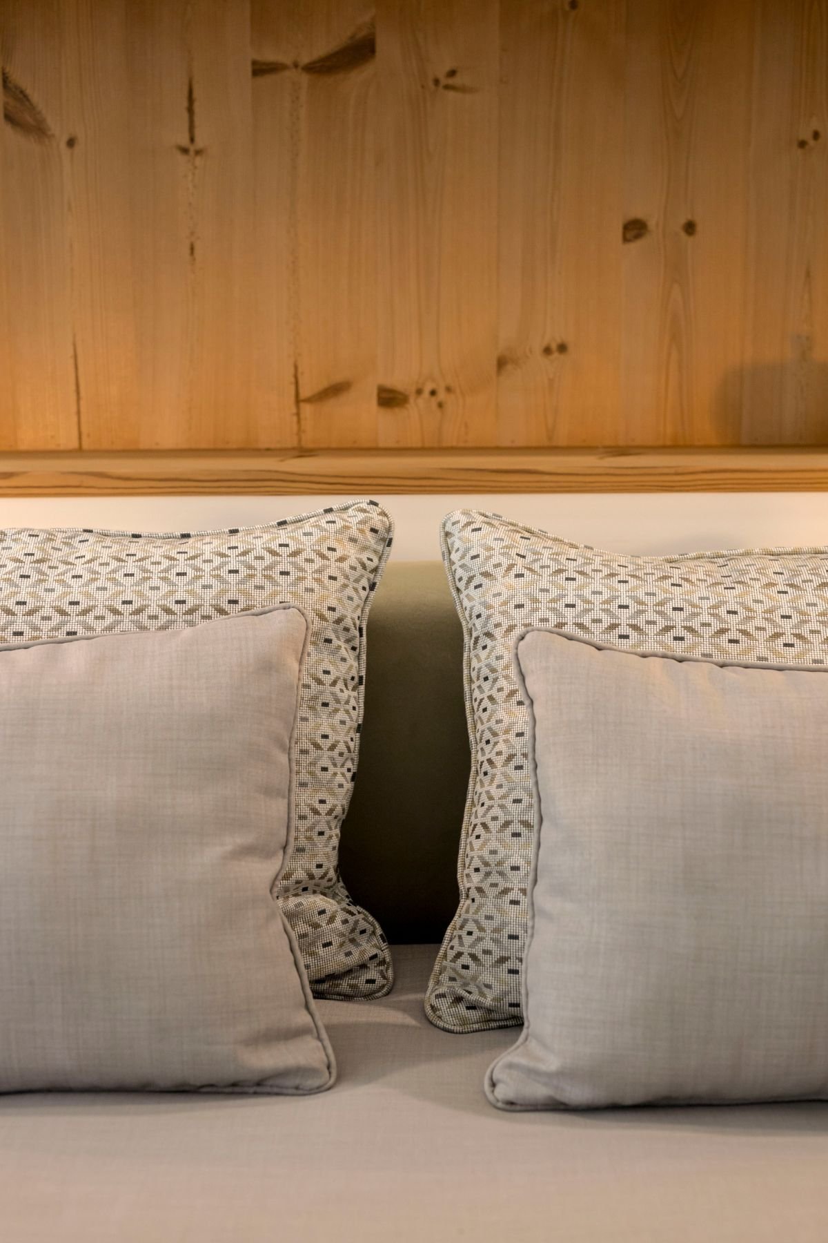 Pormenor do enxoval da cama em tons neutros e cinza, sob a textura do painel/nicho de madeira.