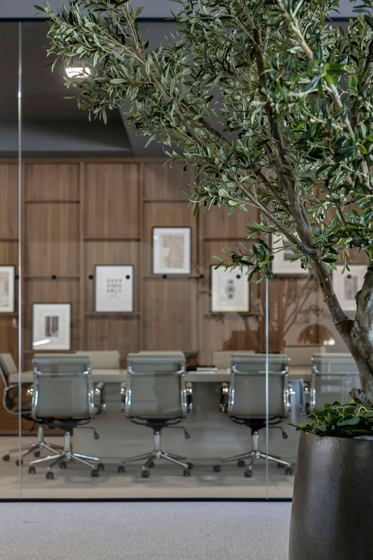 Sala de reuniões envidraçada com cadeira de escritório e painéis de madeira ao fundo vistos através de quadros decorativos.