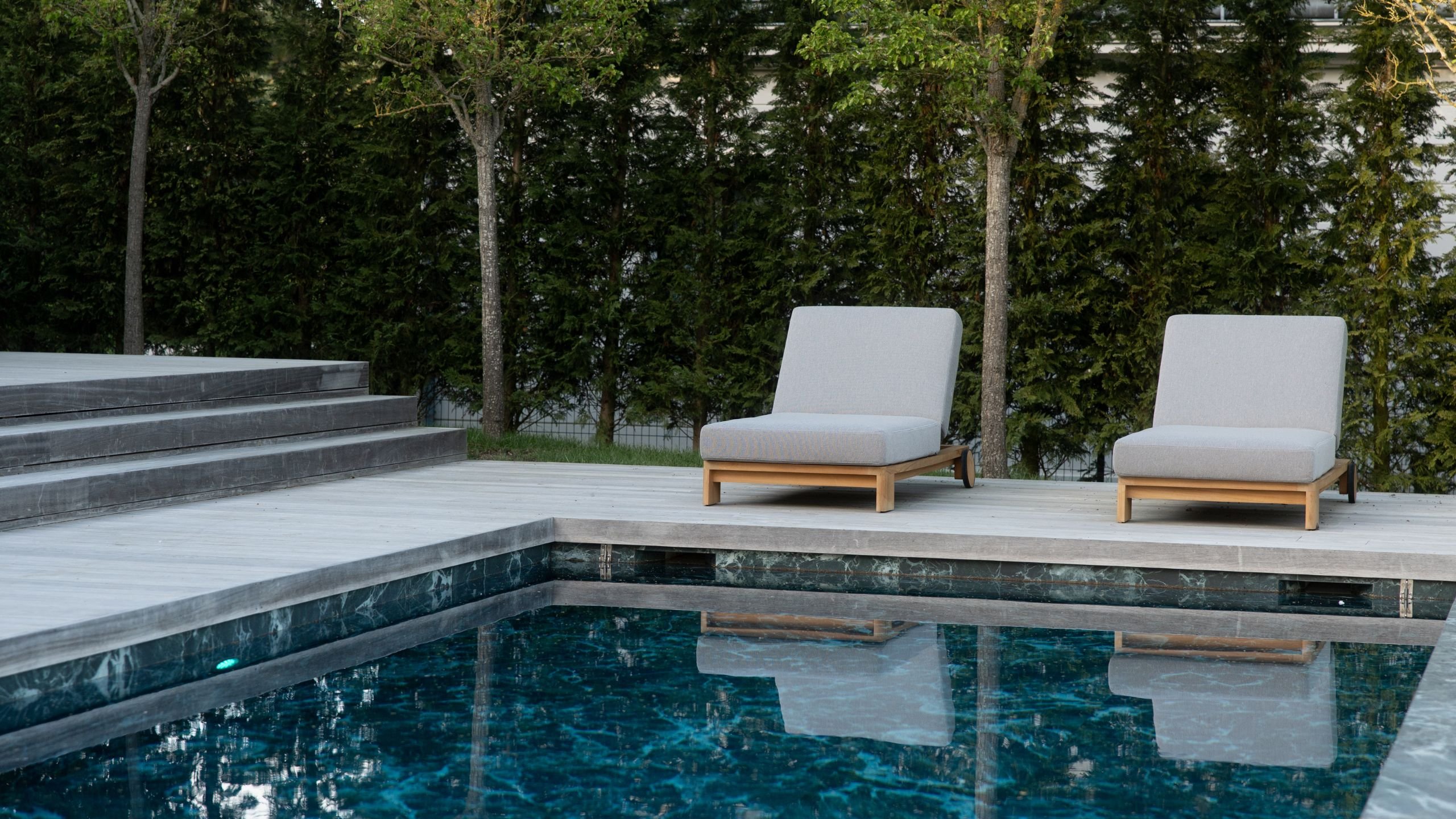 Espreguiçadeiras de exterior posicionadas no deck de madeira junto à piscina com vista para a natureza.
