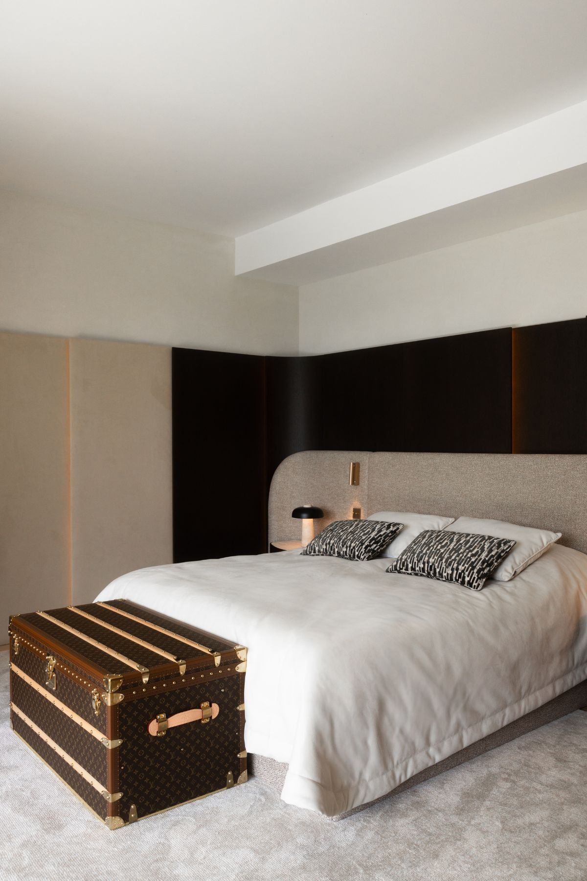 Decoração de quarto na suíte principal com cama de casal estofada, baú aos pés da cama e iluminação quente.