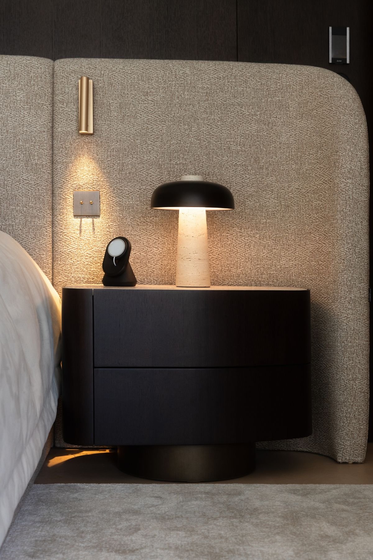 Pormenor da mesa de cabeceira curva em madeira escura com candeeiro de mesa minimalista.
