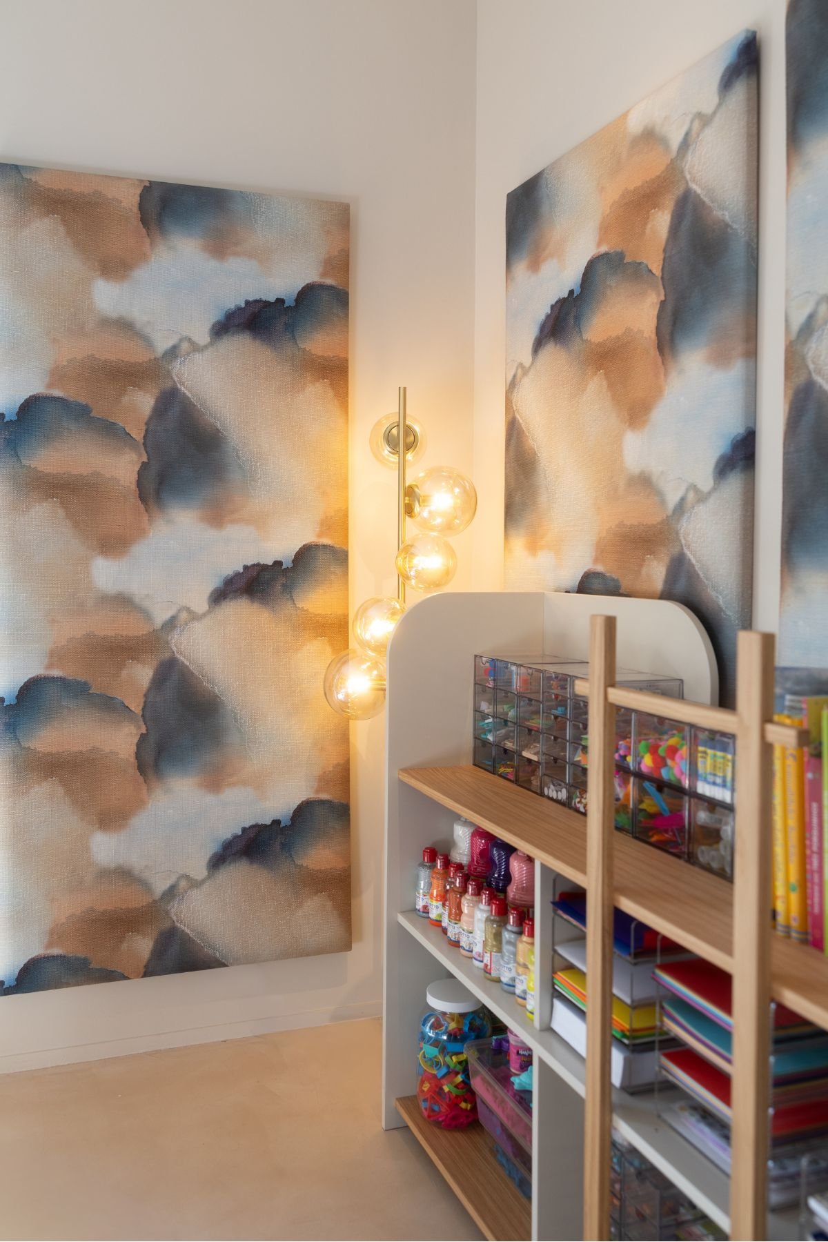 Estante de organização infantil com materiais de artes, iluminada por um aplique de parede com globos de vidro e painéis artísticos ao fundo.