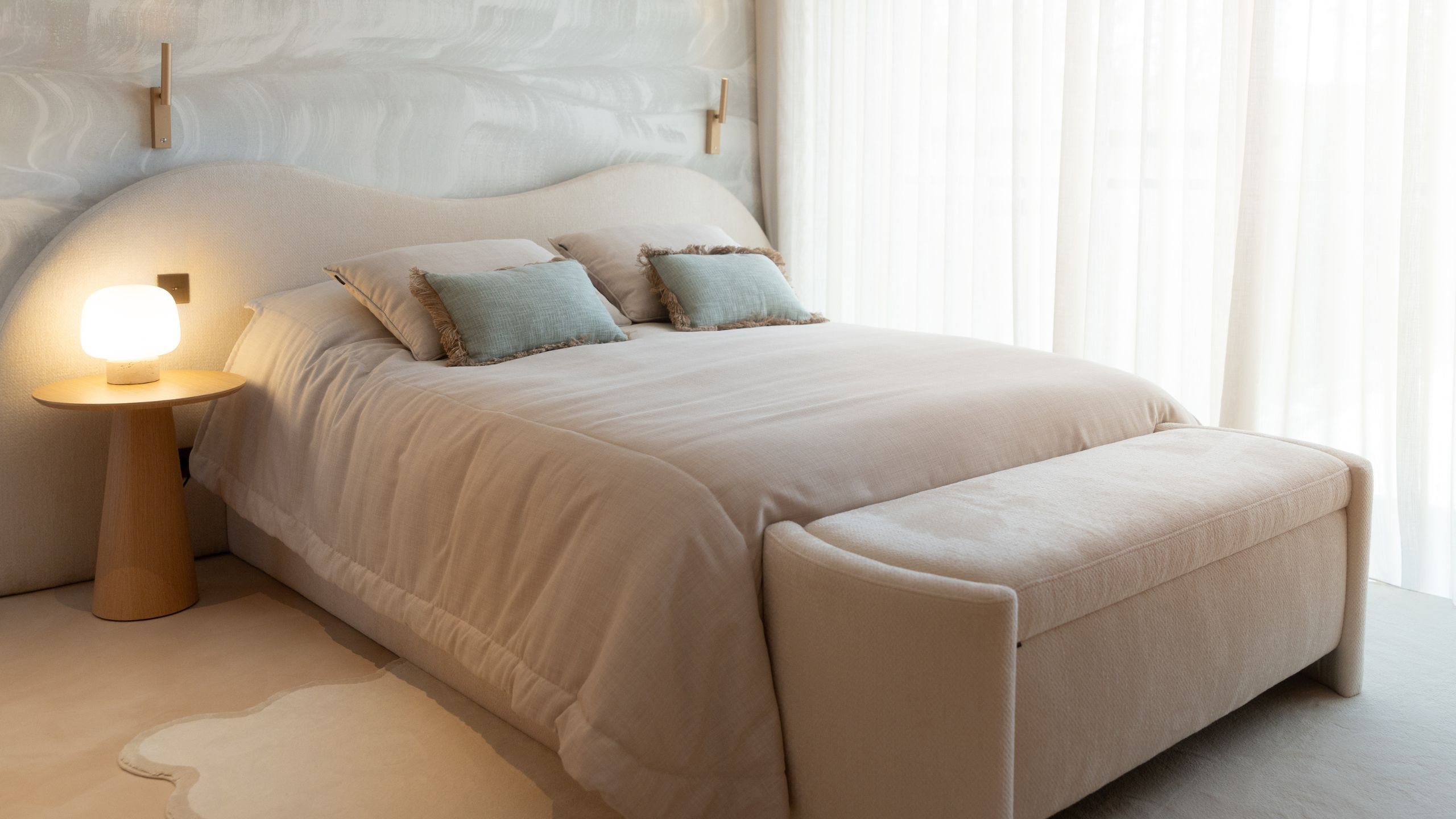 Cama de casal estofada com banco aos pés da cama, mesa de cabeceira em madeira e iluminação suave e acolhedora.