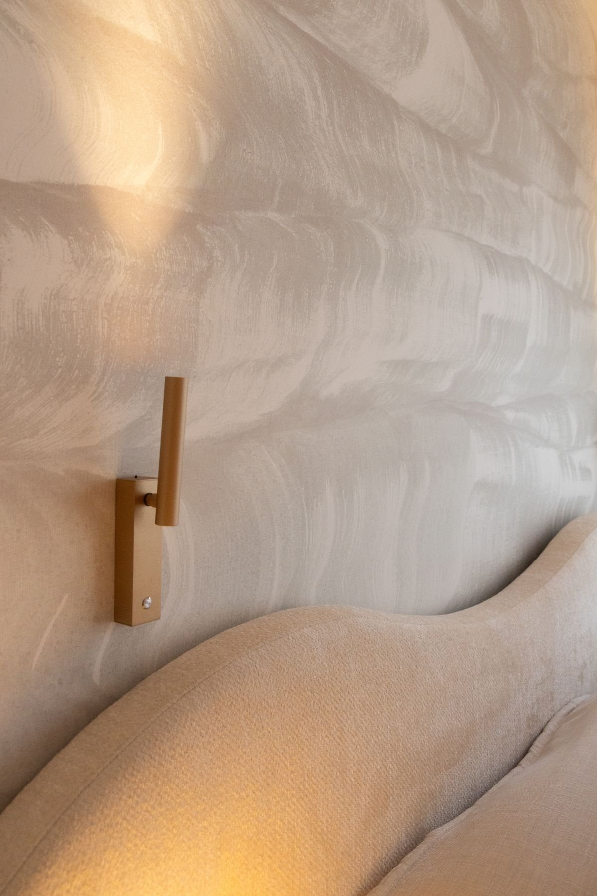 Pormenor do aplique de parede dourado e da cabeceira de cama de casal com linhas orgânicas e onduladas.