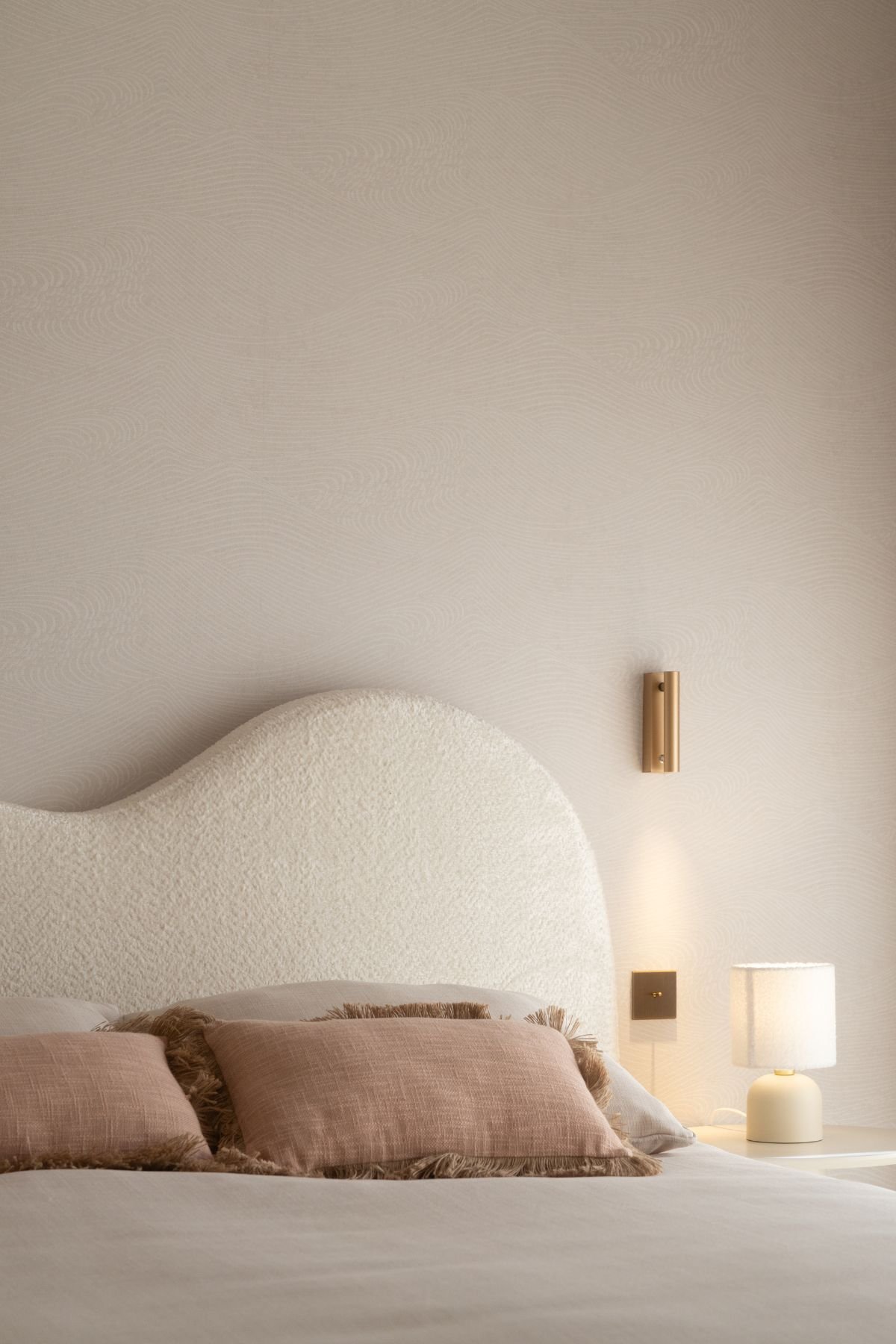 Pormenor da cama com cabeceira curva estofada e almofadas em tons rosa-seco.