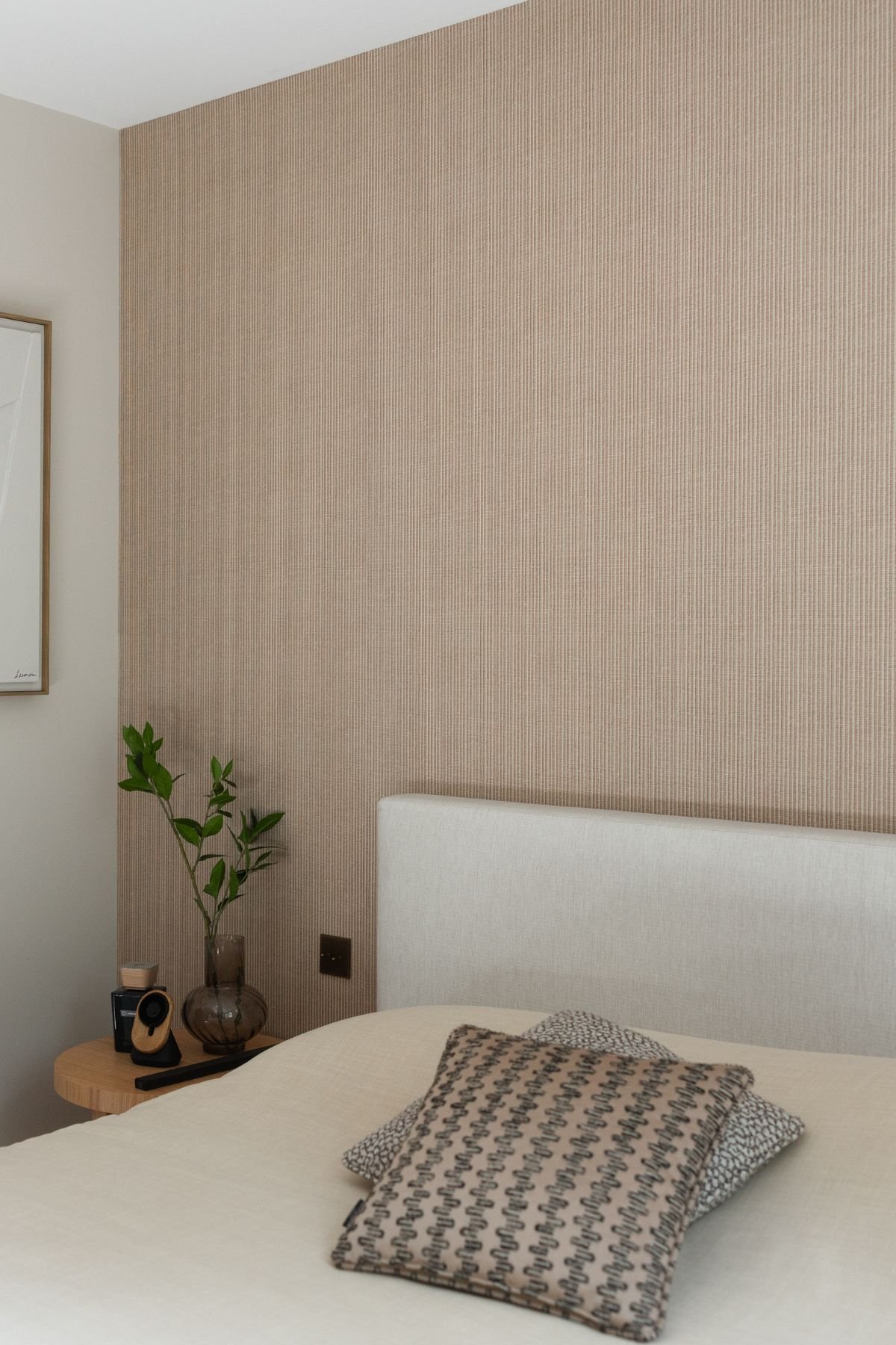 Pormenor da cama de casal com cabeceira estofada clara, mesa de cabeceira redonda com jarro decorativo e parede revestida a papel de parede textural.
