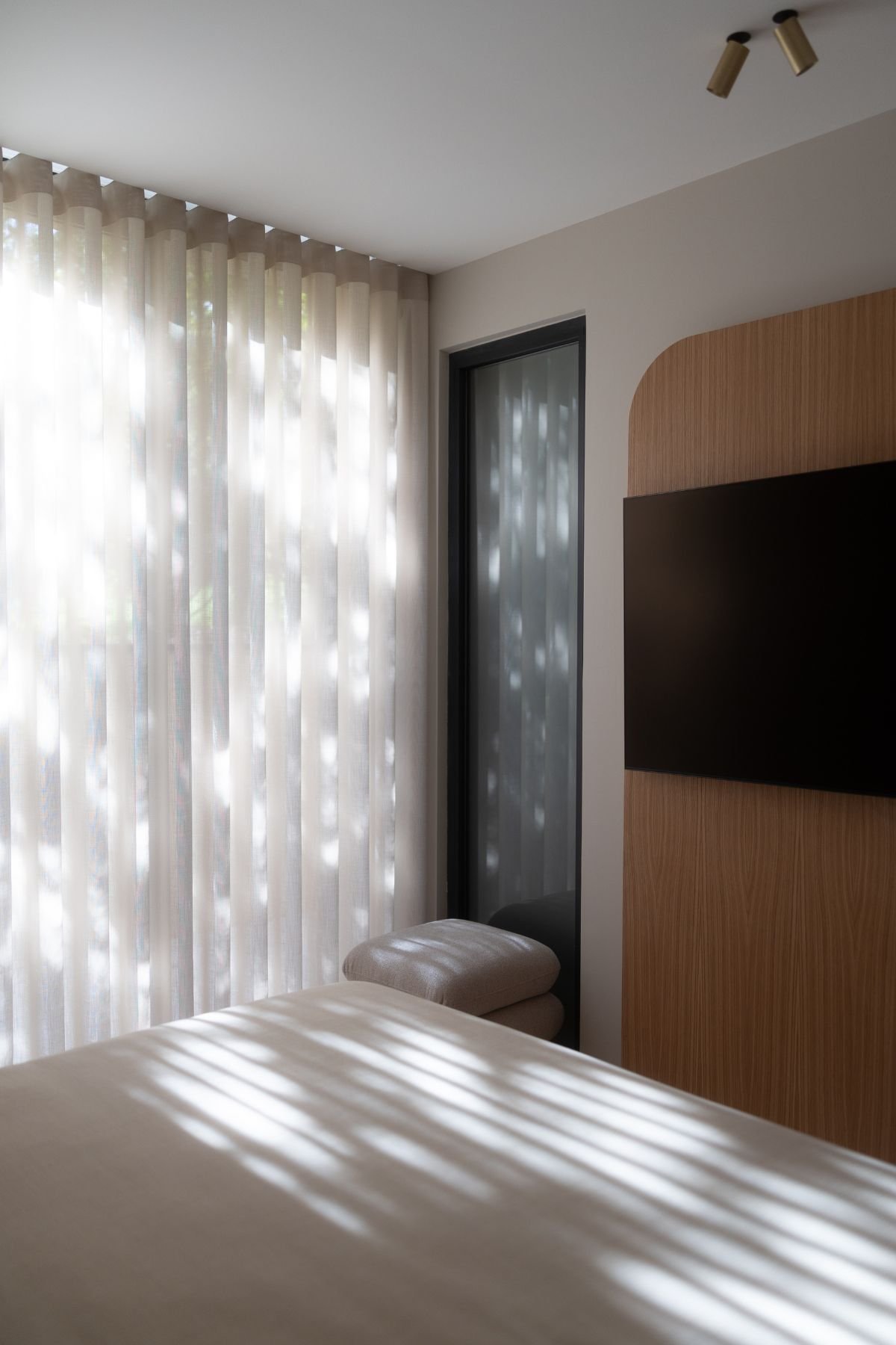 Decoração de quarto de hóspedes com jogo de luz e sombras sobre a cama de casal, cortinados translúcidos e painel de TV em madeira.