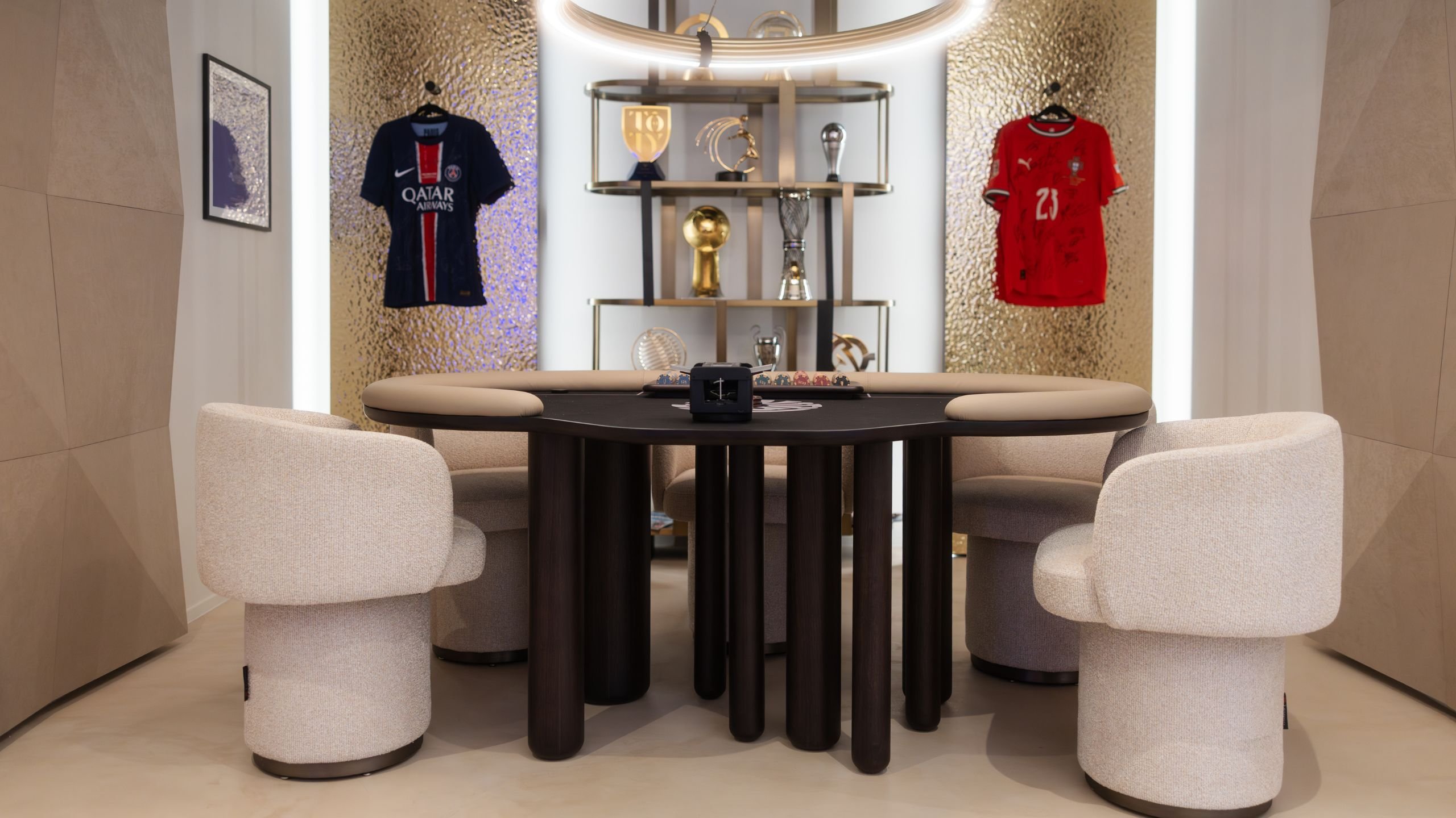 Vista geral da sala de jogos com mesa de poker personalizada, cadeiras de jantar estofadas, estante de sala com troféus e painéis em latão com camisolas enquadradas.