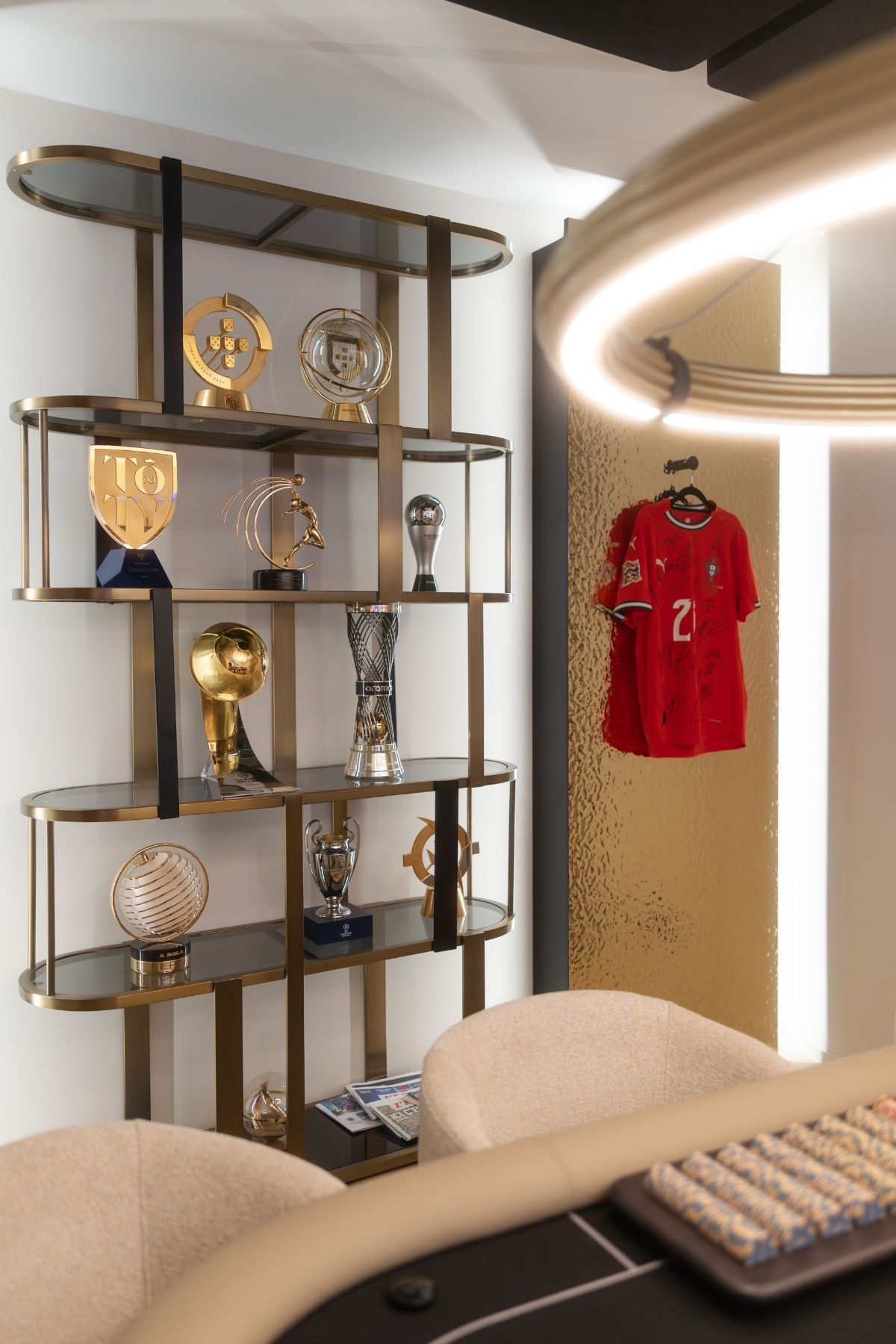 Estante de sala metálica em tom dourado com prateleiras em vidro a exibir troféus e painéis de parede decorativos com camisolas de futebol.