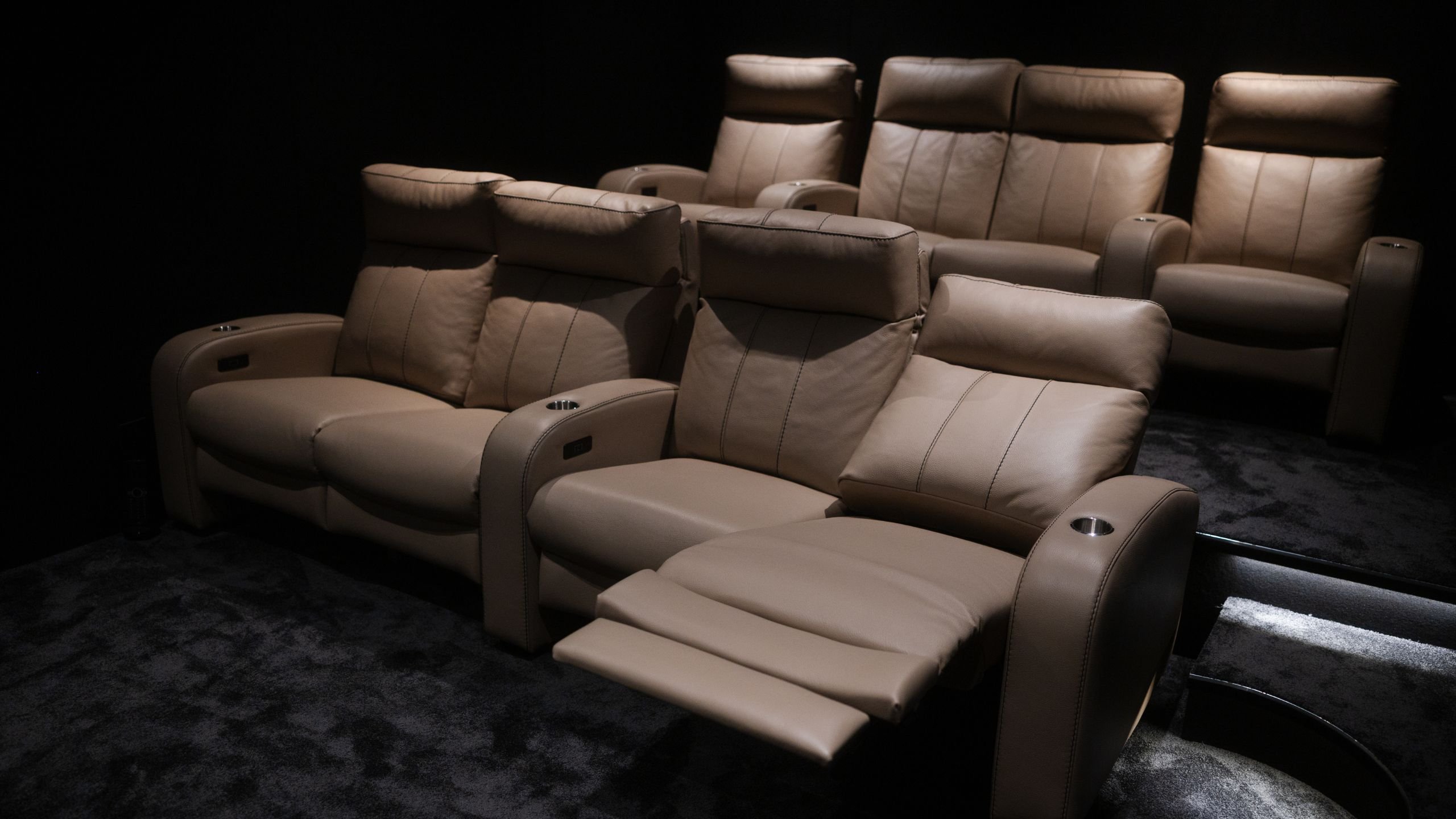 Perspetiva das filas de poltronas reclináveis em pele da sala de cinema em casa, com piso alcatifado e degraus iluminados.
