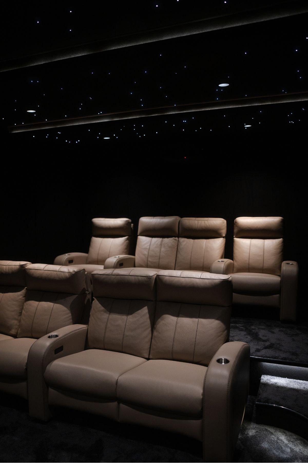 Vista geral da sala de cinema privada com poltronas reclináveis em pele bege, dispostas em anfiteatro, sob um teto estrelado.