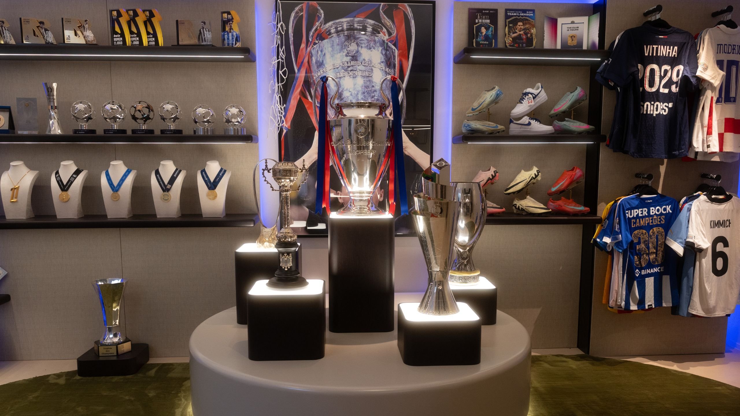 Vista frontal da zona de exposição com camisolas de futebol penduradas, troféus em pedestais iluminados e iluminação técnica.