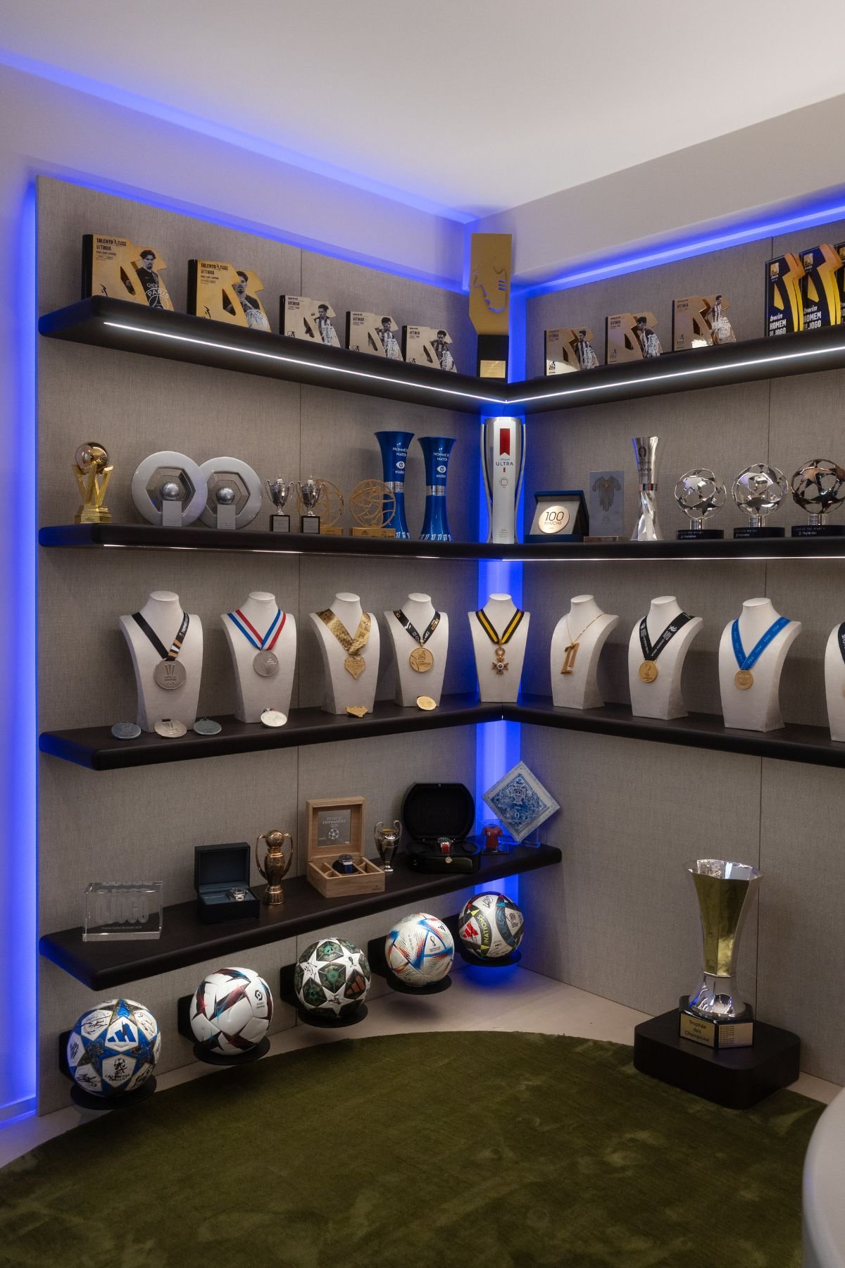 Prateleiras retroiluminadas com iluminação LED azul a expor medálias, bolas de futebol e prémios individuais.