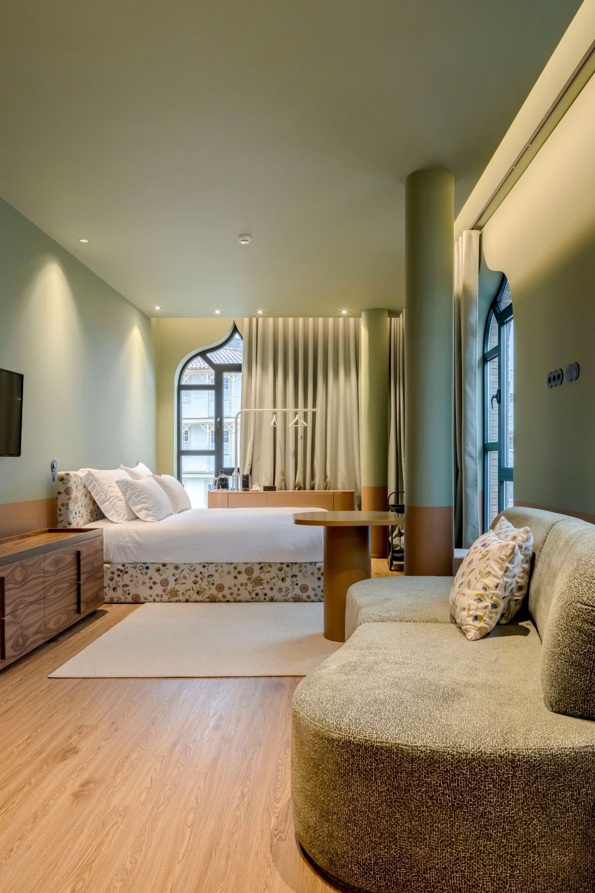 Perspetiva ampla do quarto integrando a zona de descanso com a cama, a área de estar com sofá e o mobiliário em madeira.