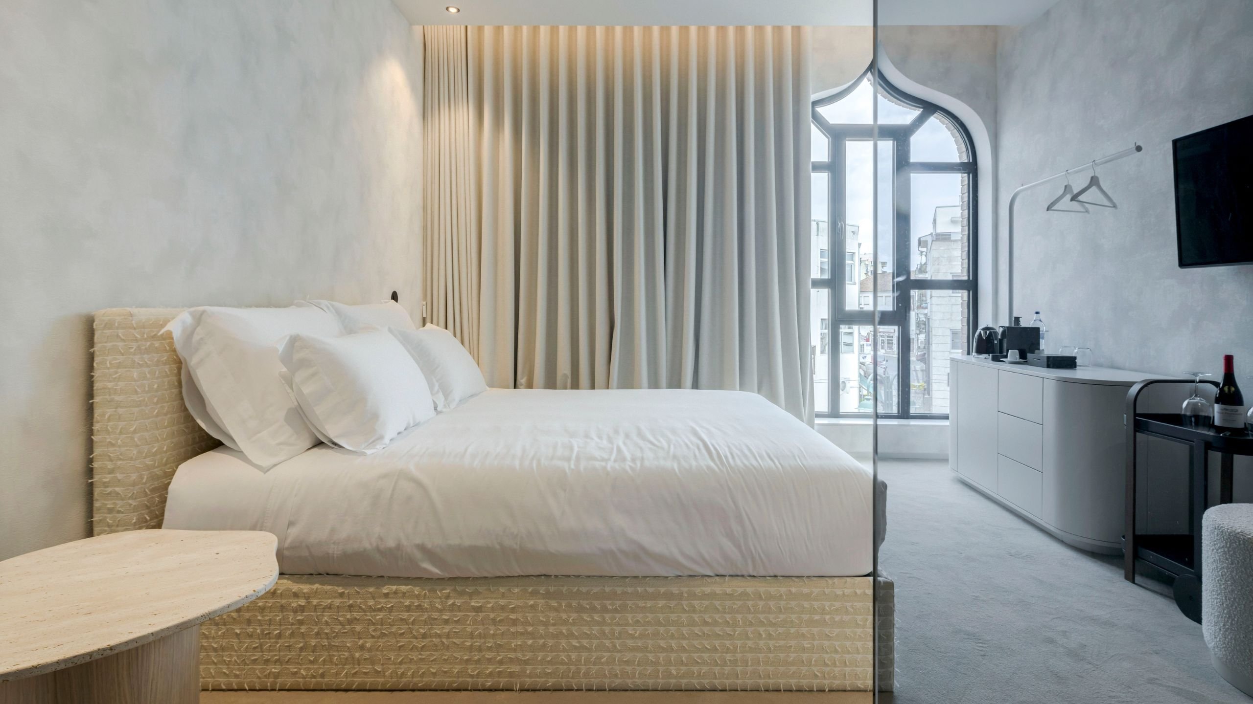Vista geral do quarto com a cama estofada em tecido fil coupé, delimitada pelo vidro fumê da casa de banho e com a mesa de apoio em destaque no primeiro plano.