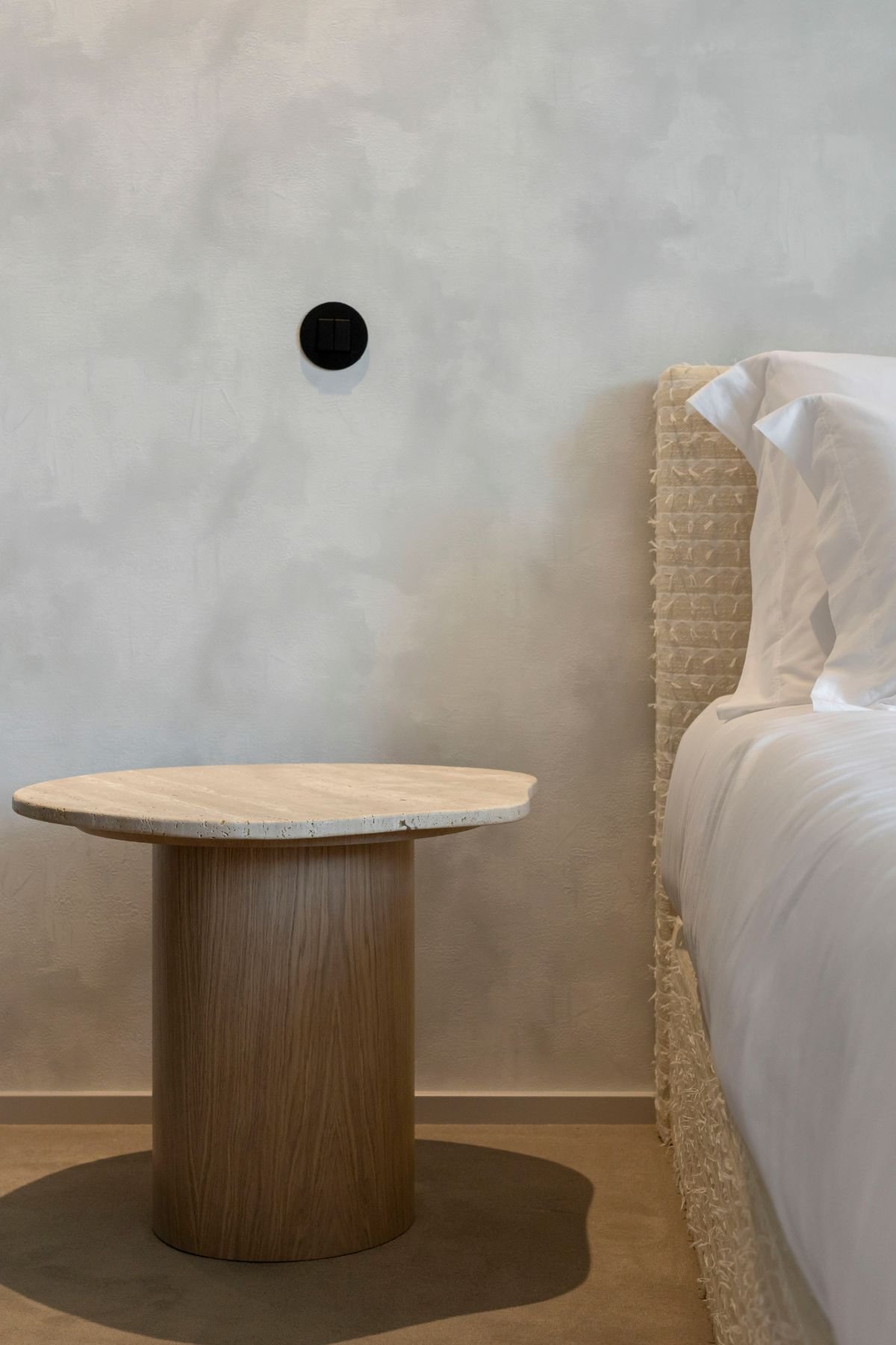 Detalhe da mesa de apoio com tampo orgânico em mármore travertino e base cilíndrica em carvalho, junto à cabeceira da cama.