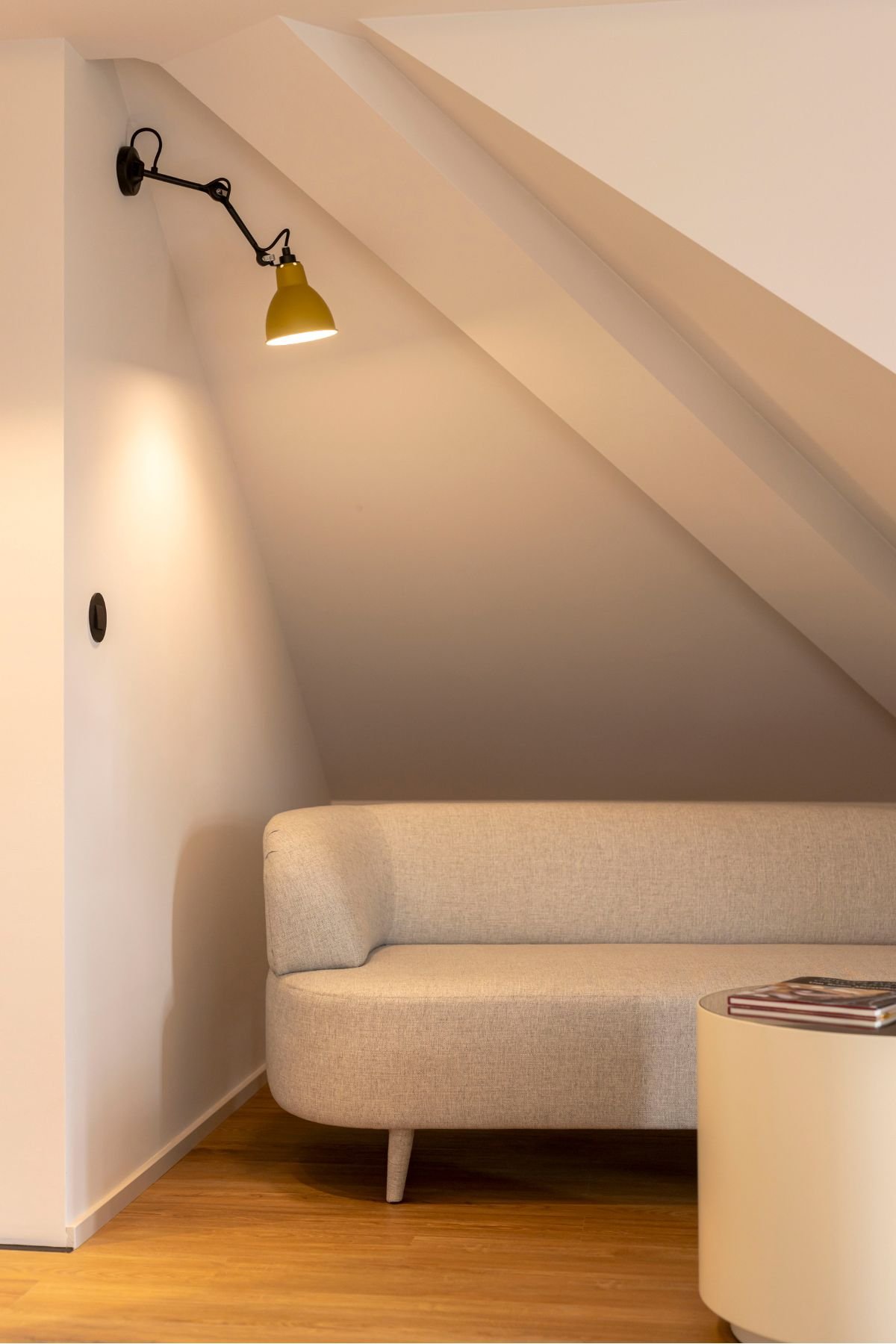 Pormenor do recanto lounge com o sofá neutro e o aplique de parede articulado amarelo sob o teto esconso.