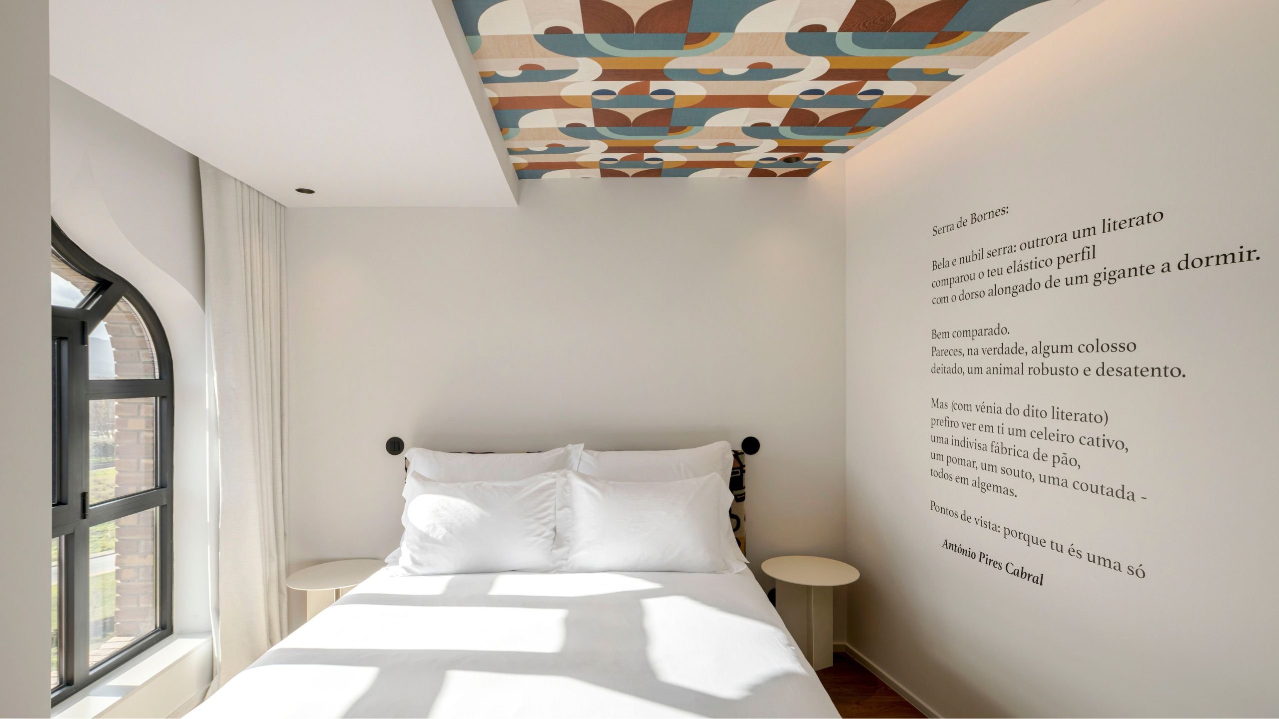 Zona da cama decorada com poesia impressa na parede principal, ladeada por mesas de cabeceira circulares brancas, papel de parede abstrato no teto e janela em arco.